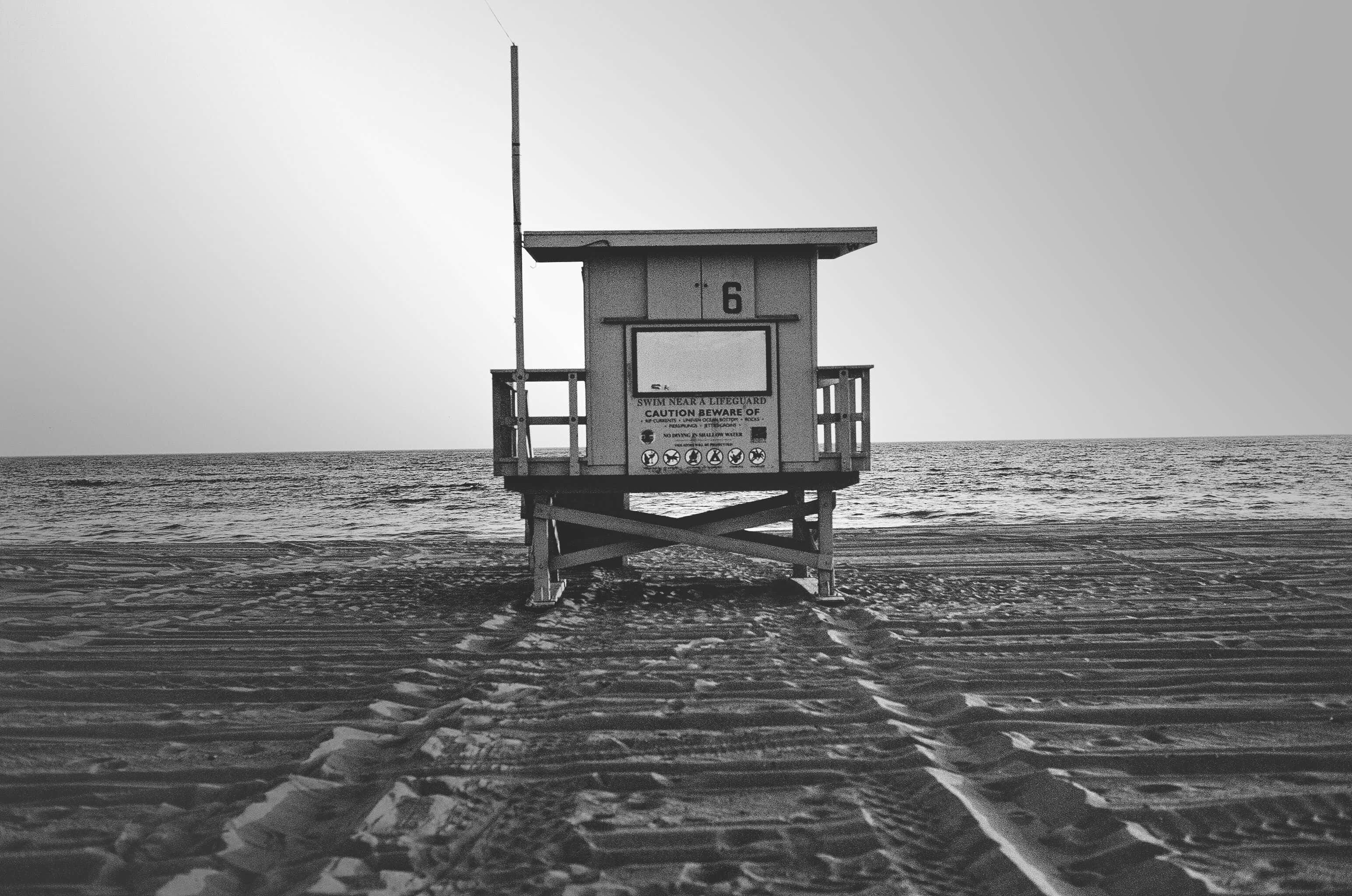 Black And White Beach 桌布 壁紙 — 一座孤獨的救生員瞭望塔矗立在紋理沙灘上，面向平靜的海洋，天空朦朧，呈現單色調。