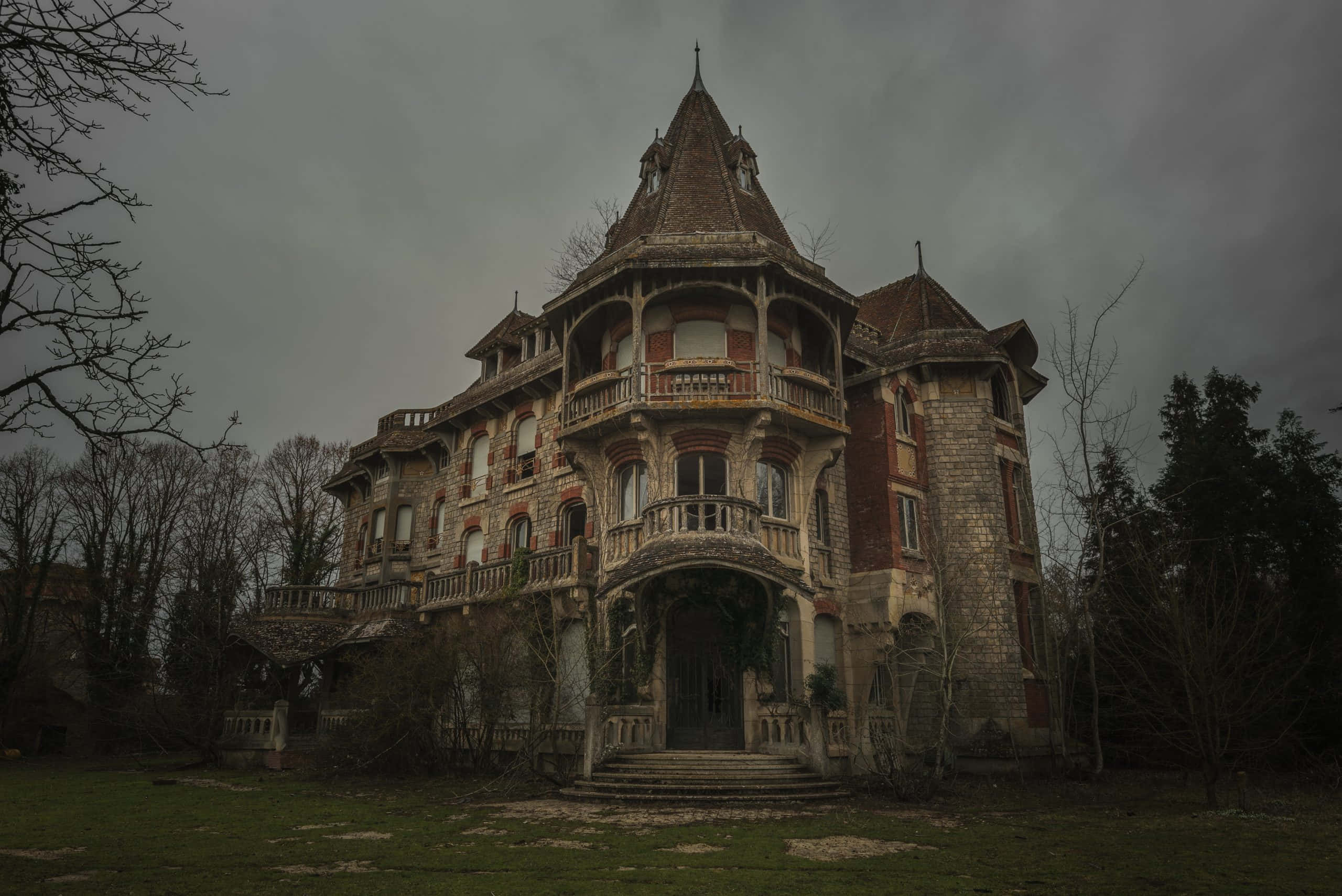 Haunted House 桌布 壁紙 — 一座宏偉但破敗的大宅，擁有華麗的陽台和石塔，矗立在陰沉多雲的天空下，周圍是光禿禿的樹木。