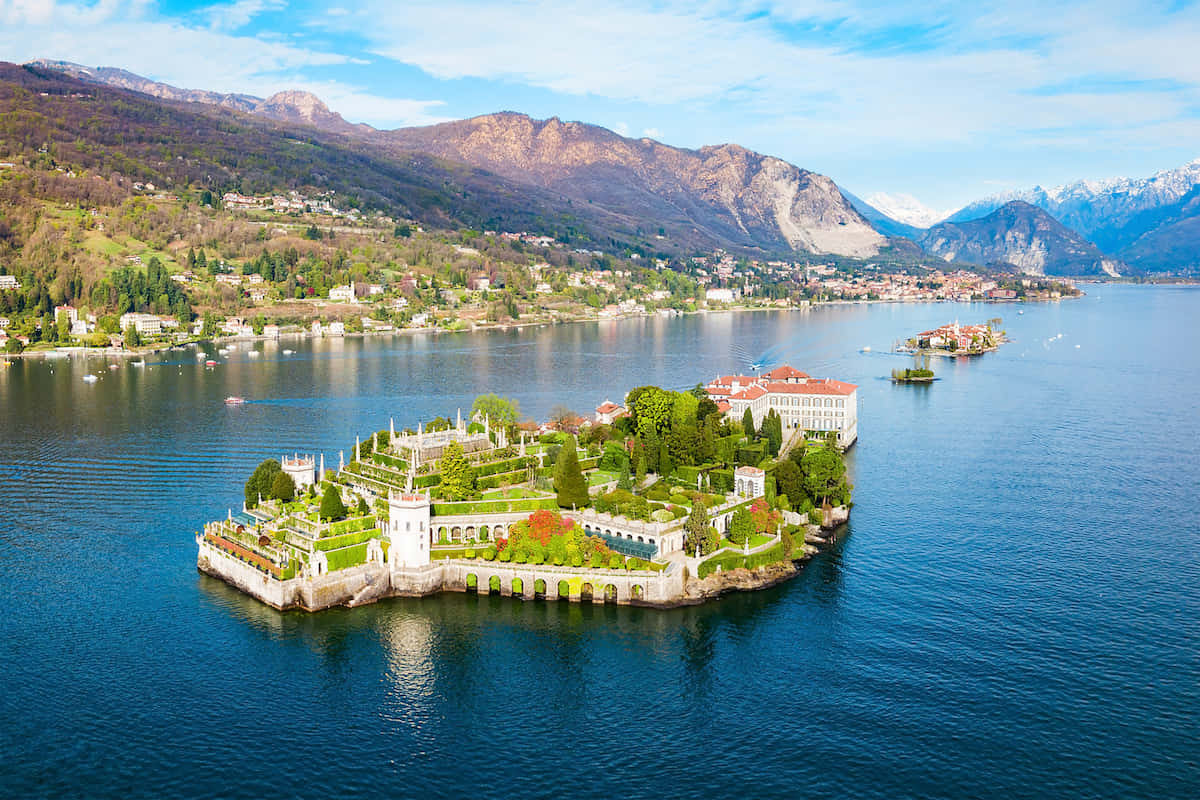 Italian Island 桌布 壁紙 — 一座宏偉島嶼別墅的鳥瞰圖，擁有修剪整齊的花園，漂浮在深藍色的水面上，周圍是森林覆蓋的山脈和白雪皚皚的山峰，在明亮的藍天下。