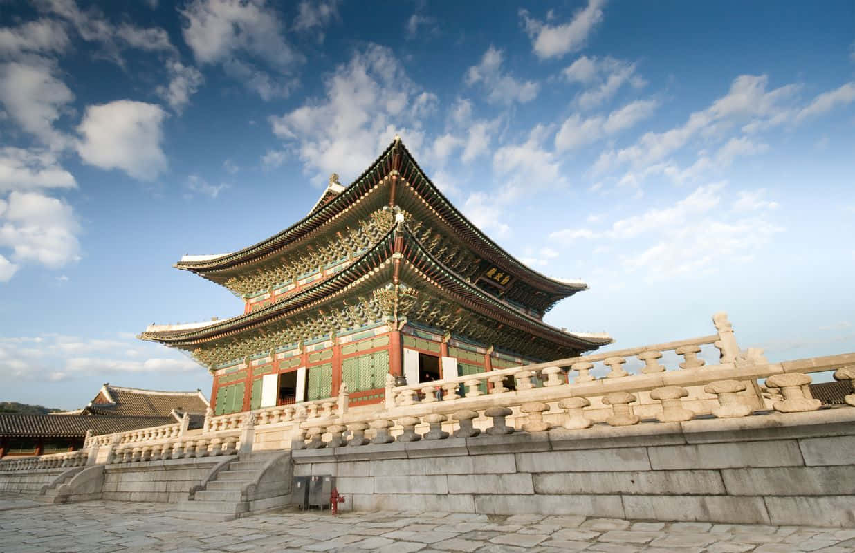 South Korea 桌布 壁紙 — 一座宏偉的傳統韓國宮殿，擁有華麗的瓦片屋頂和紅綠色的牆壁，在明亮的藍天和蓬鬆的白雲下。