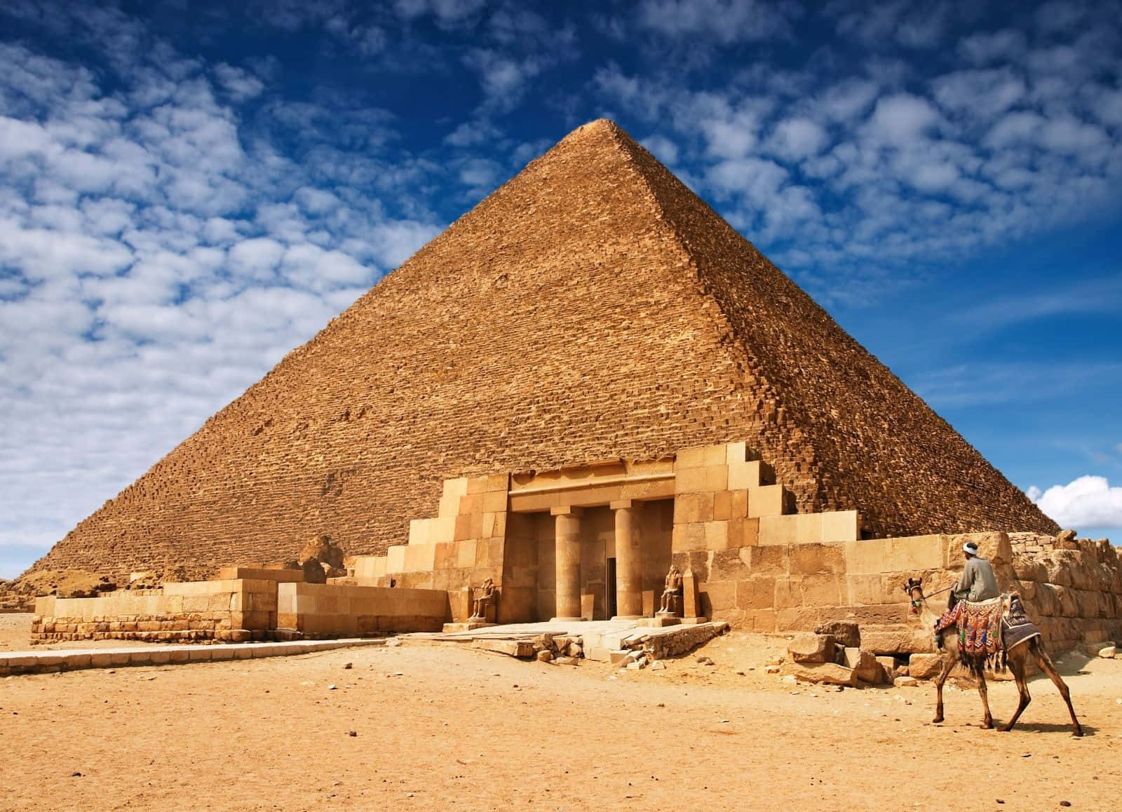 Egypt 桌布 壁紙 — 一座宏偉的古埃及金字塔在點綴著蓬鬆白雲的鮮豔藍天下聳立，一位騎駱駝的旅人正穿越金色的沙漠。