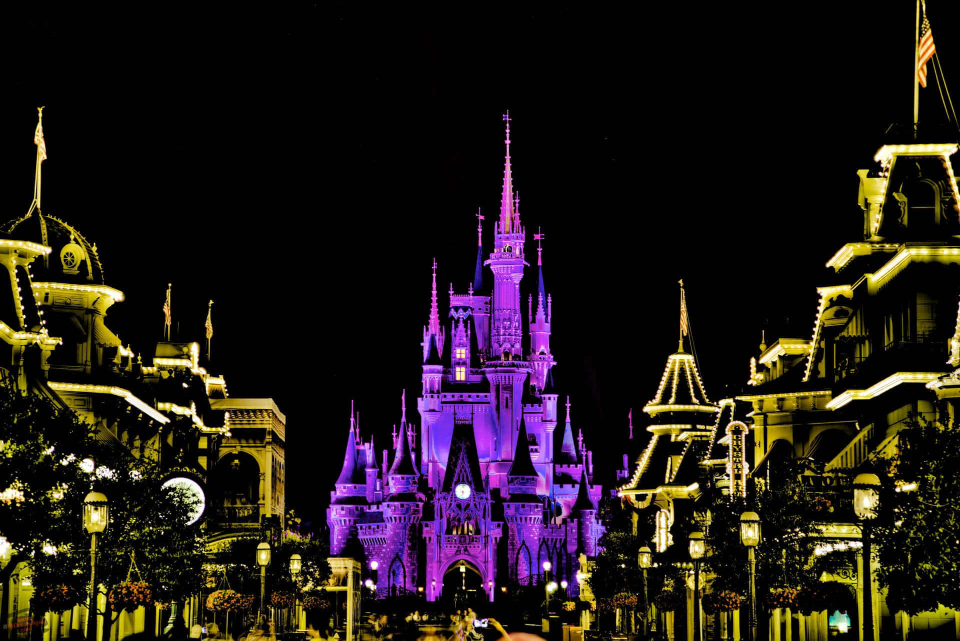 Disney Castle 桌布 壁紙 — 一座宏偉的城堡在夜間被鮮豔的紫色燈光照亮，矗立於中央，兩旁是溫暖燈光的建築物，背景是漆黑的天空。