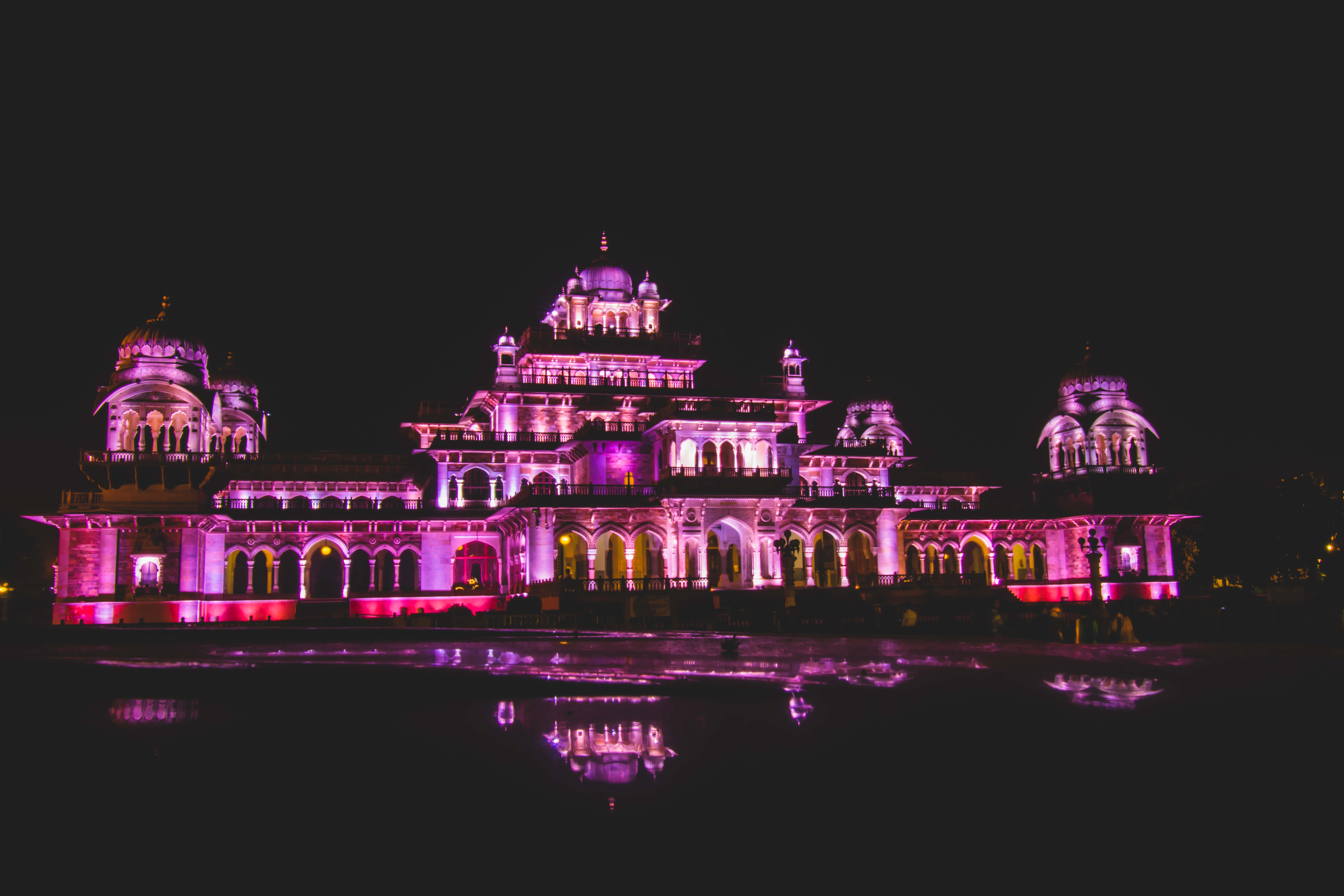 Rajasthan 桌布 壁紙 — 一座宏偉的宮殿在夜晚被鮮豔的粉紅色和紫色燈光照亮，美麗地倒映在平靜的水面上。