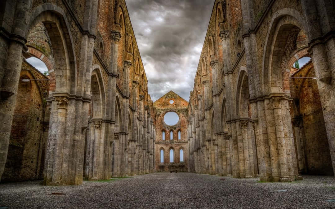 Gothic Architecture 桌布 壁紙 — 一座宏偉的廢棄修道院，高聳的石拱門和石柱矗立在戲劇性的多雲天空下。