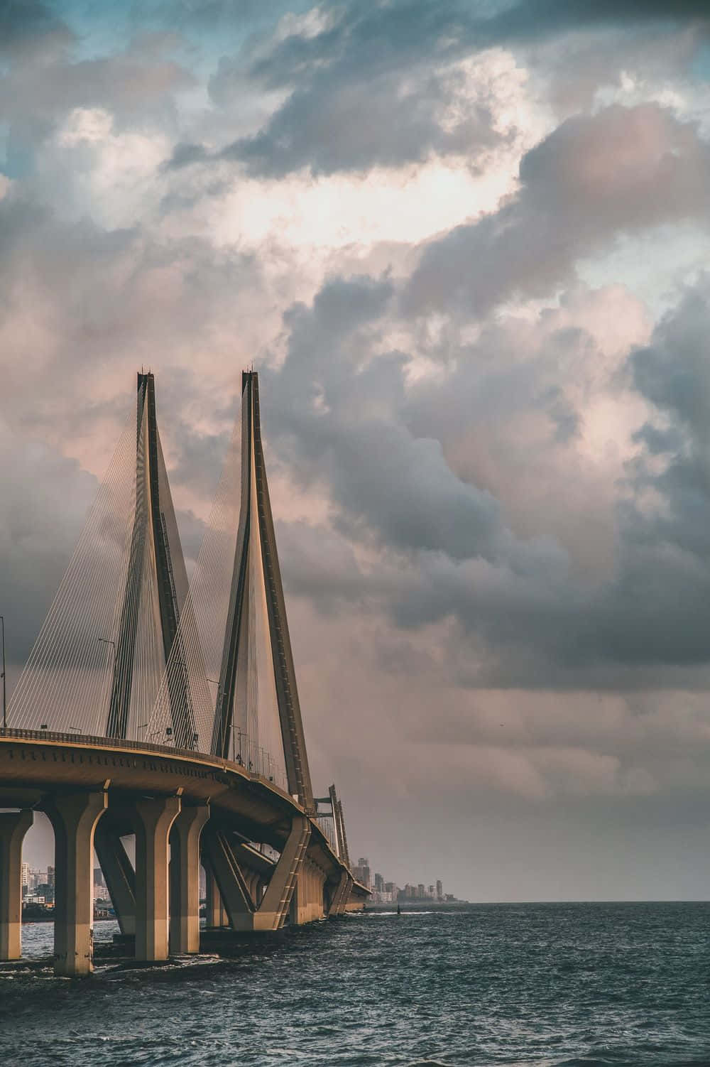 Mumbai 桌布 壁紙 — 一座宏偉的斜拉橋在黃昏戲劇性的多雲天空下延伸在平靜的水面上。