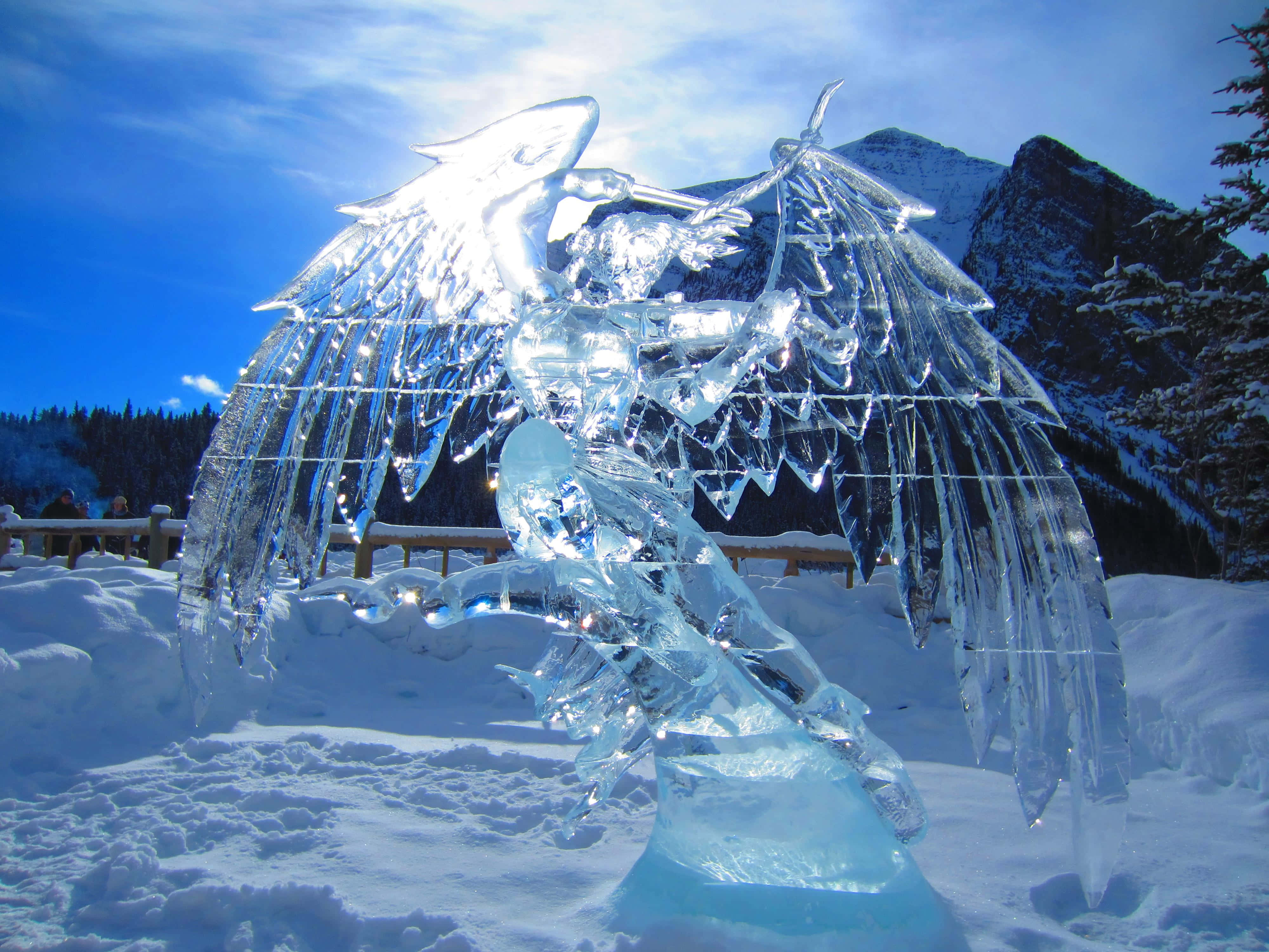 Ice Sculpture 桌布 壁紙 — 一座宏偉的有翼生物冰雕矗立在白雪皚皚的山景中，天空湛藍明亮。