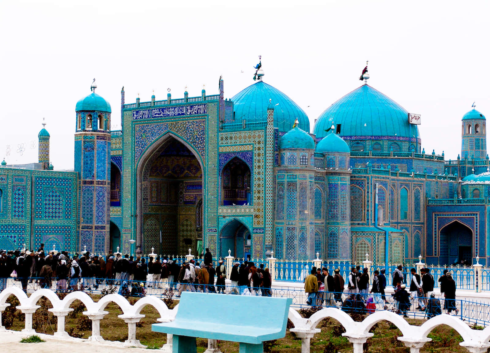 Afghanistan 桌布 壁紙 — 一座宏偉的清真寺，擁有充滿活力的藍綠色圓頂和複雜的瓷磚工藝，矗立在多雲的天空下，人群聚集在白色拱形圍欄附近。