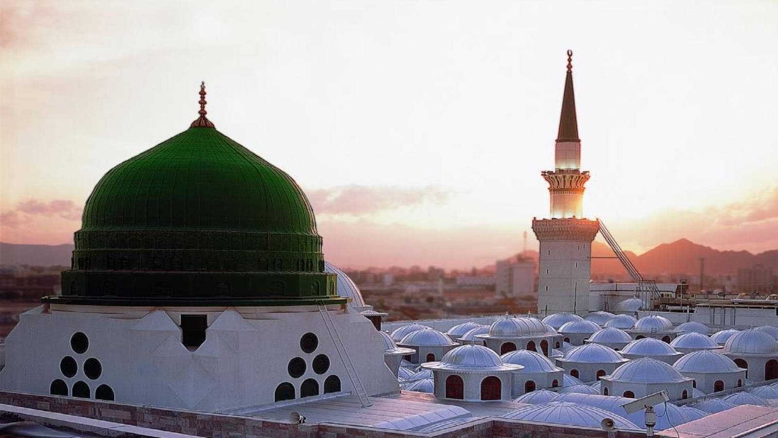 Madina Sharif 桌布 壁紙 — 一座宏偉的清真寺，擁有充滿活力的綠色圓頂和高聳的宣禮塔，在金色的夕陽下沐浴，背景是朦朧的天空。