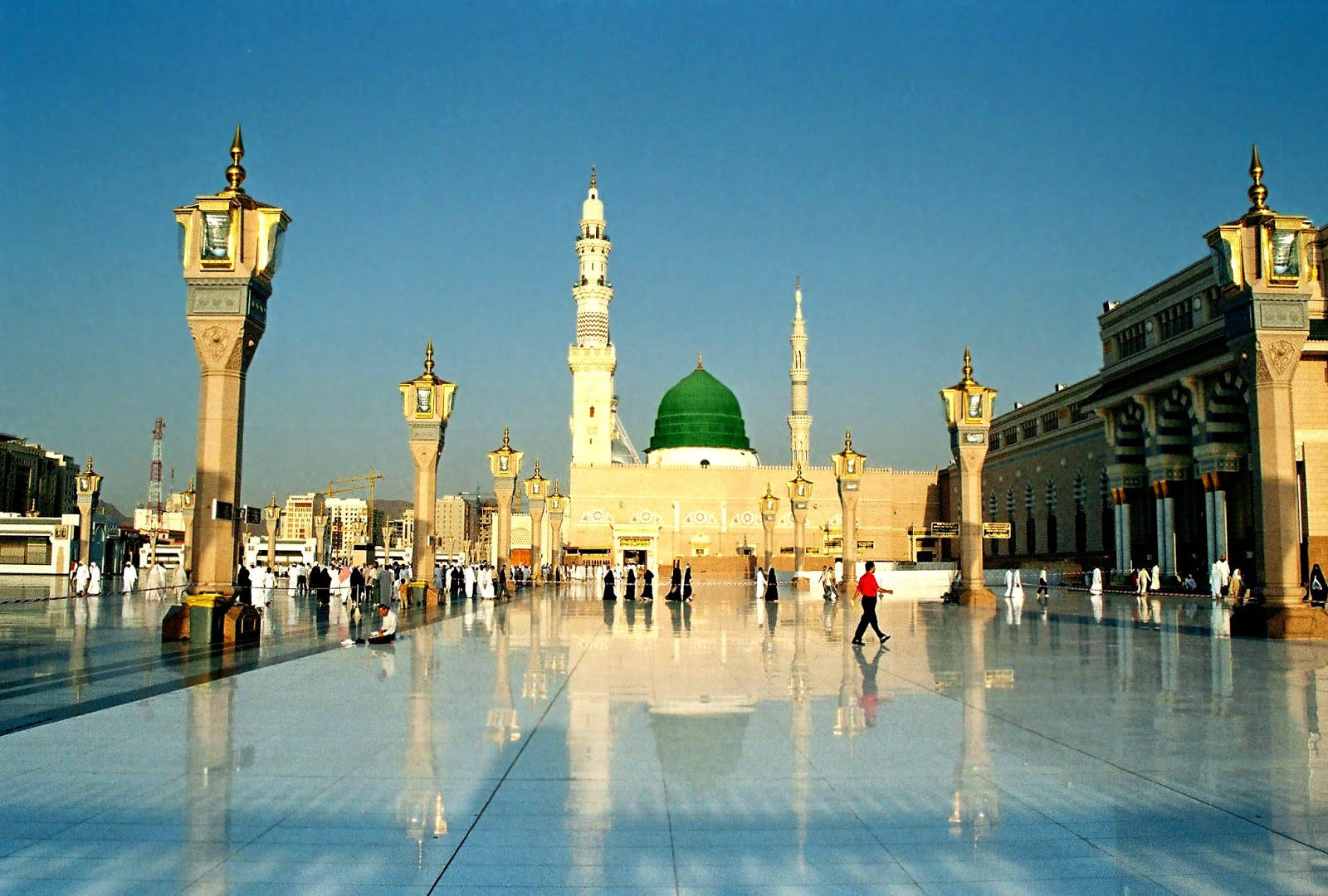 Madina Sharif 桌布 壁紙 — 一座宏偉的清真寺，擁有綠色圓頂和高聳的宣禮塔，矗立在清澈的藍天下，倒映在人們行走的廣闊拋光廣場上。