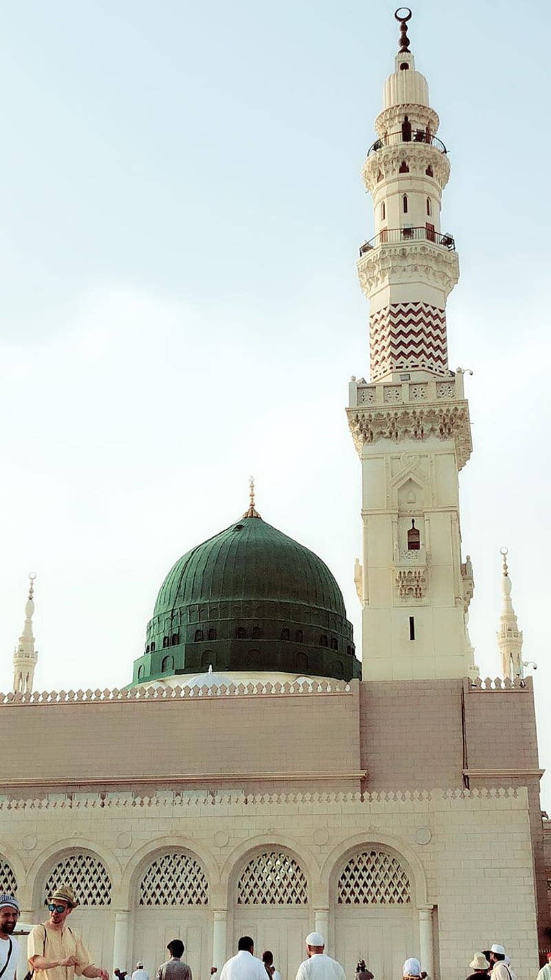Madina Sharif 桌布 壁紙 — 一座宏偉的清真寺，擁有綠色圓頂和高聳的宣禮塔，矗立在蒼白的天空下，信徒們聚集在它的底部。