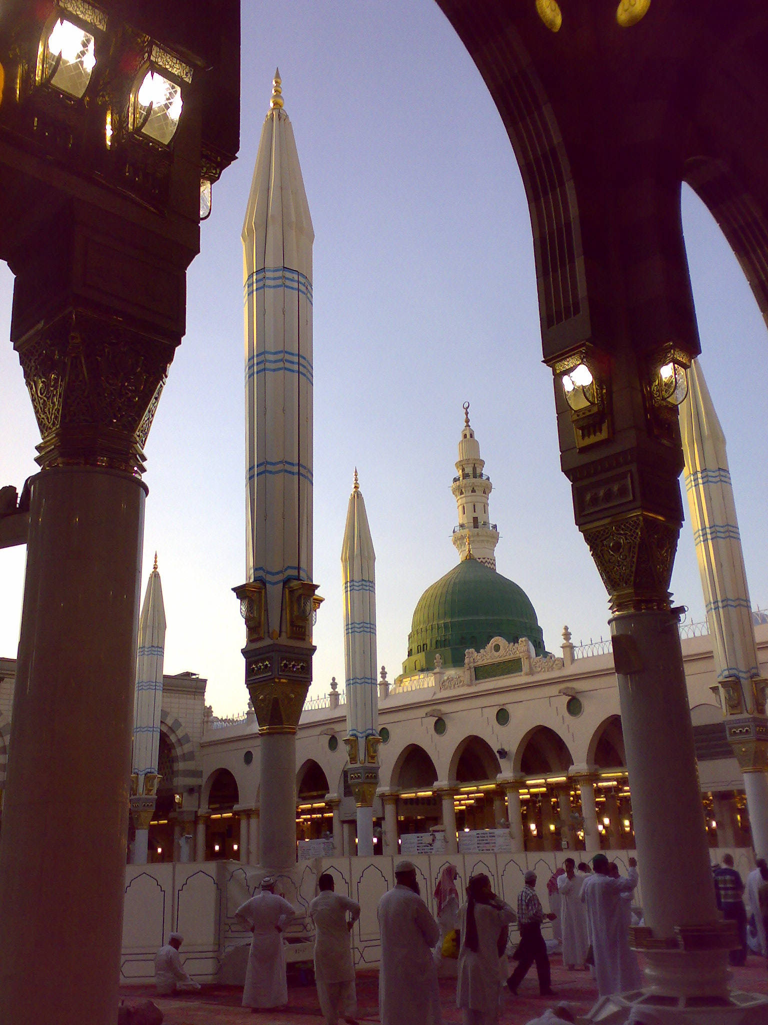 Madina Sharif 桌布 壁紙 — 一座宏偉的清真寺，擁有綠色圓頂和高聳的宣禮塔，透過華麗的拱門觀看，信徒們在黃昏時聚集。