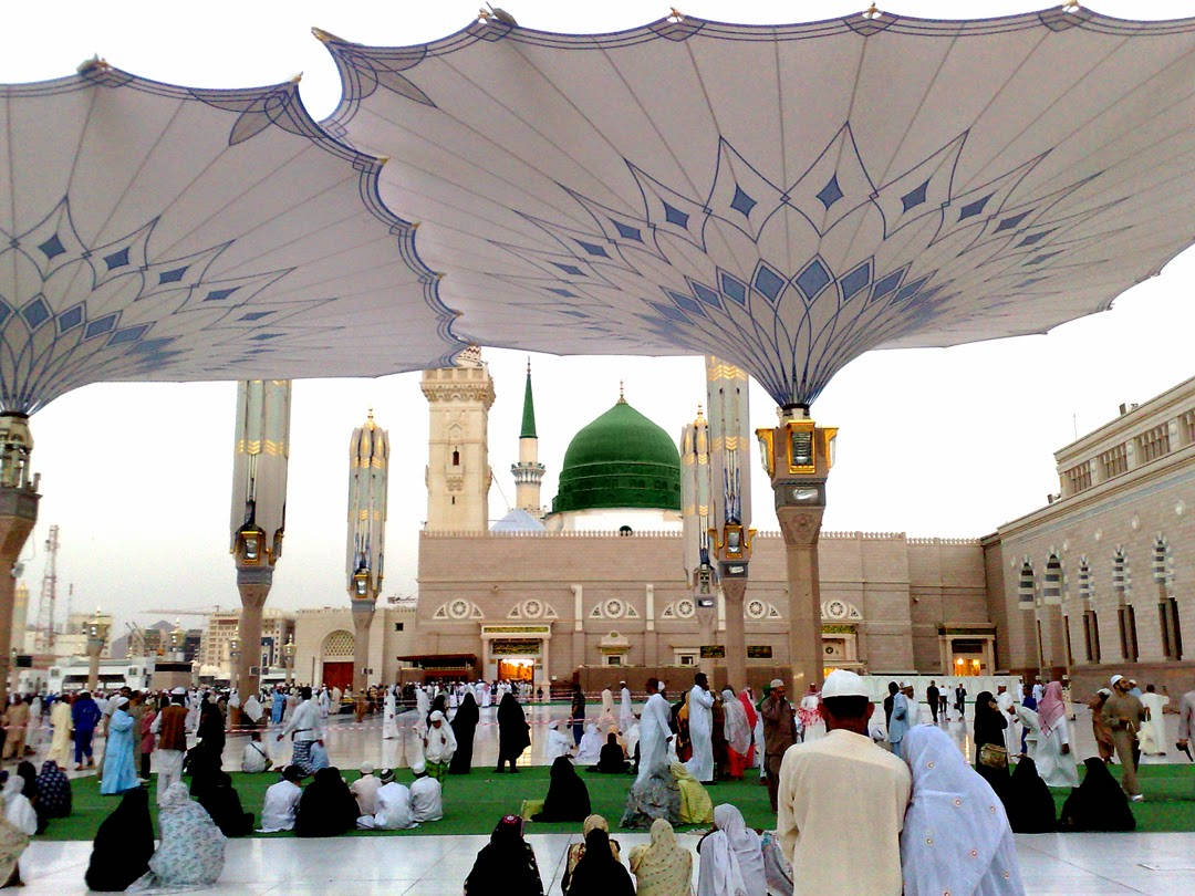 Madina Sharif 桌布 壁紙 — 一座宏偉的清真寺，擁有綠色圓頂和高聳的宣禮塔，矗立於大型圖案天篷下，信徒們聚集在寬敞的庭院中。
