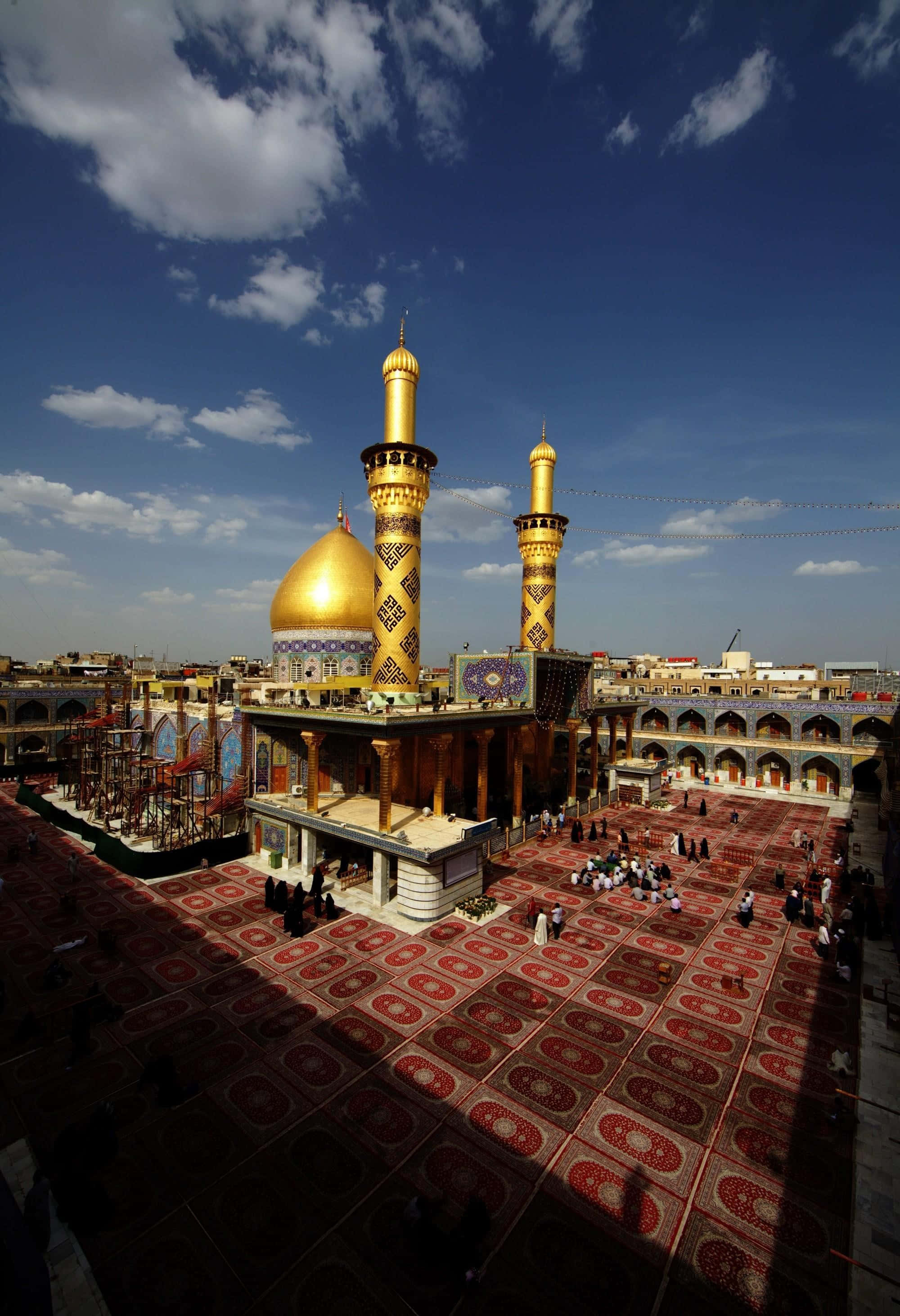 Karbala 桌布 壁紙 — 一座宏偉的清真寺，擁有金色圓頂和宣禮塔，矗立在明亮的藍天下，散落的雲朵在紅色圖案祈禱毯上投下陰影。