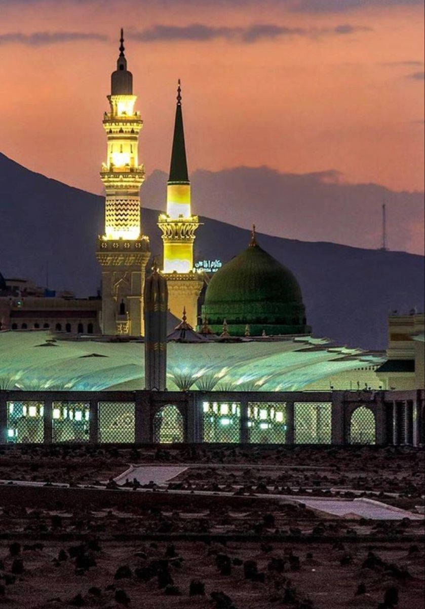 Madina Sharif 桌布 壁紙 — 一座宏偉的清真寺，發光的宣禮塔和綠色圓頂，矗立在火紅的日落天空下，背後是剪影般的山脈。