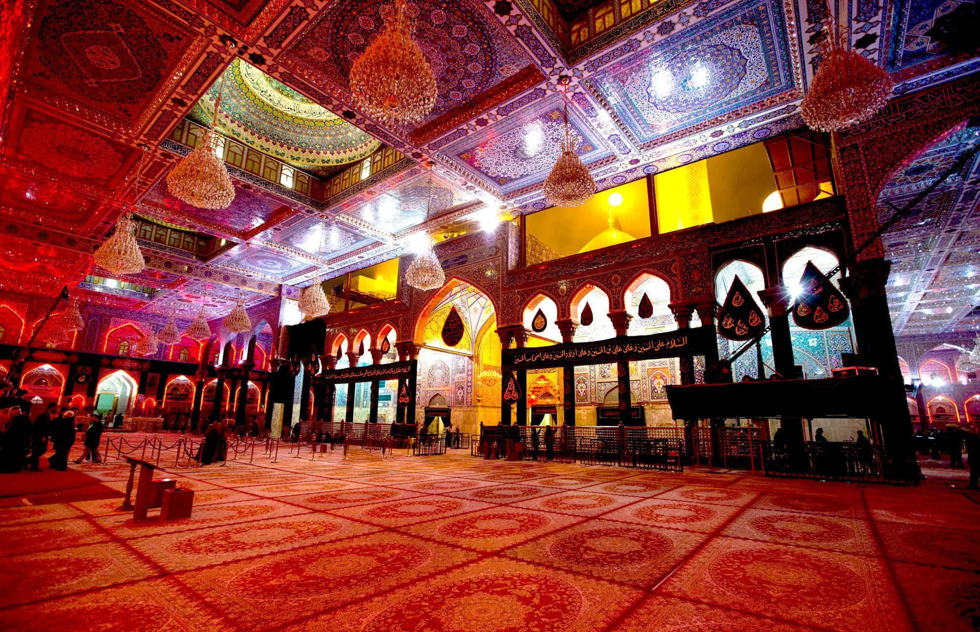 Karbala 桌布 壁紙 — 一座宏偉的清真寺內部，擁有華麗的紅金色瓷磚地板、複雜的天花板圖案和懸掛的吊燈，在溫暖的人造燈光下。