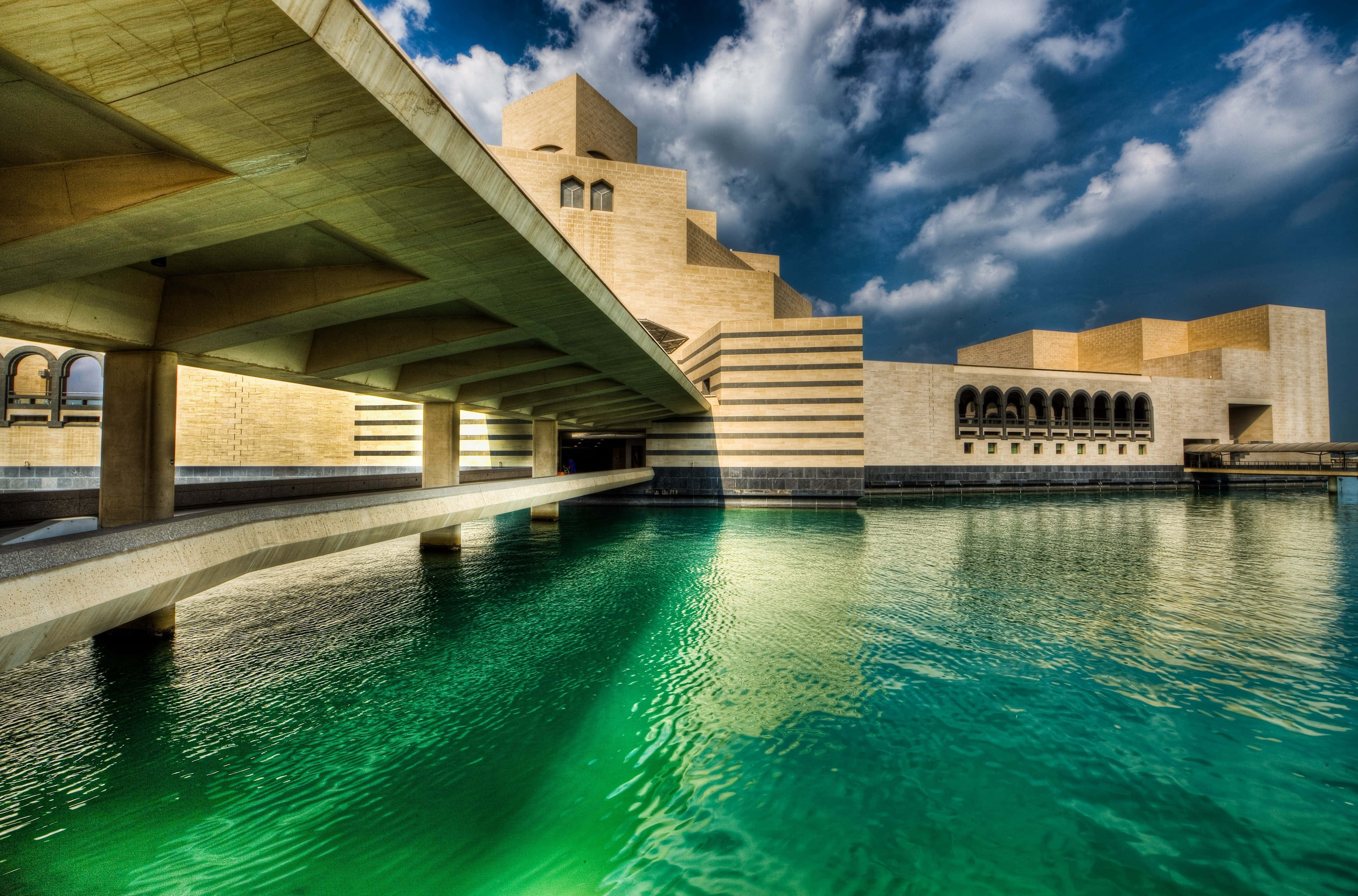 Museums 桌布 壁紙 — 一座宏偉的現代主義建築，帶有幾何石塊，矗立在充滿活力的綠色水域旁，天空戲劇性地多雲。