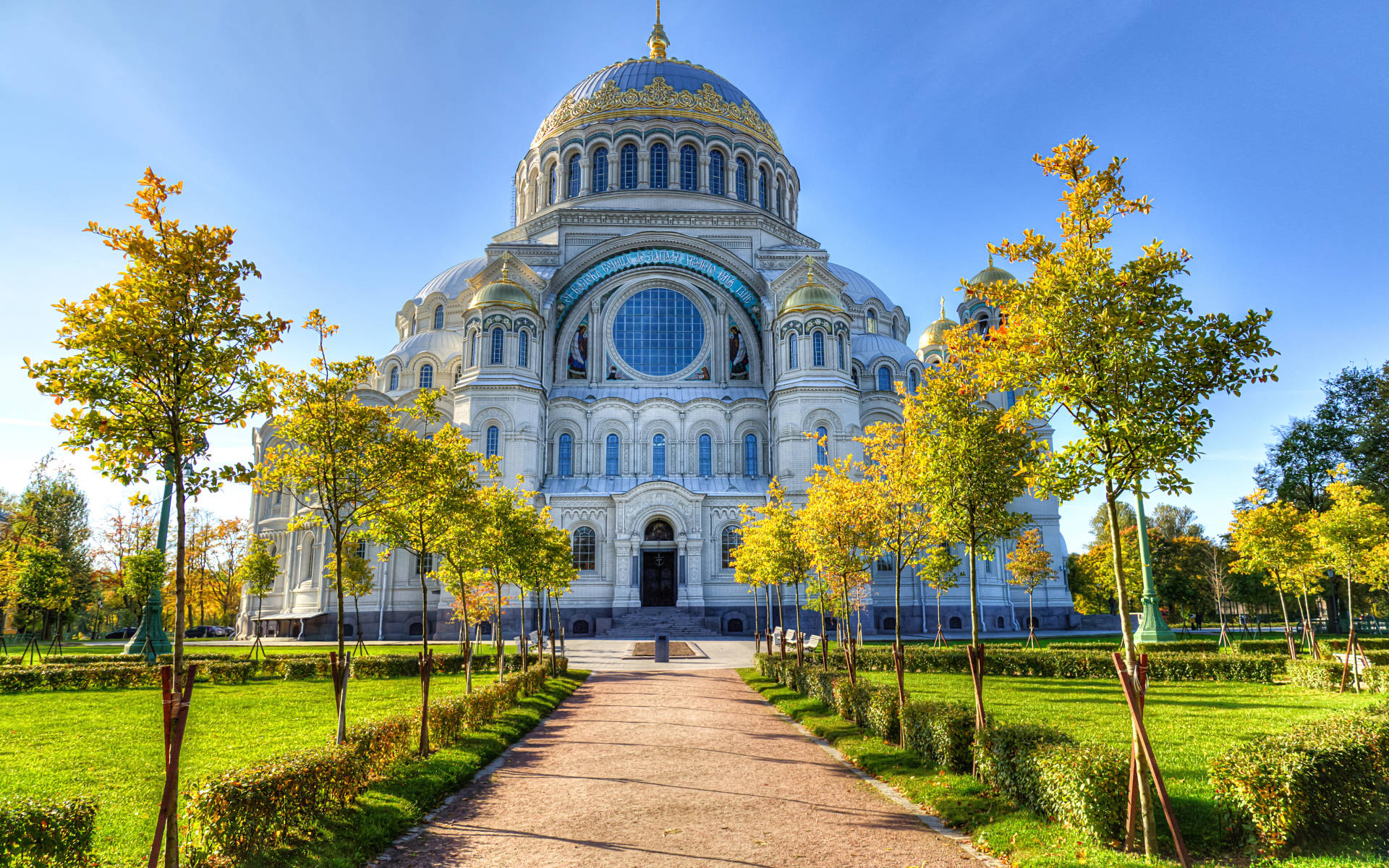 Russia 桌布 壁紙 — 一座宏偉的白色大教堂，擁有金色圓頂，矗立在清澈的藍天下，周圍是秋日黃葉的樹木，一條鋪砌的小徑通往其入口。