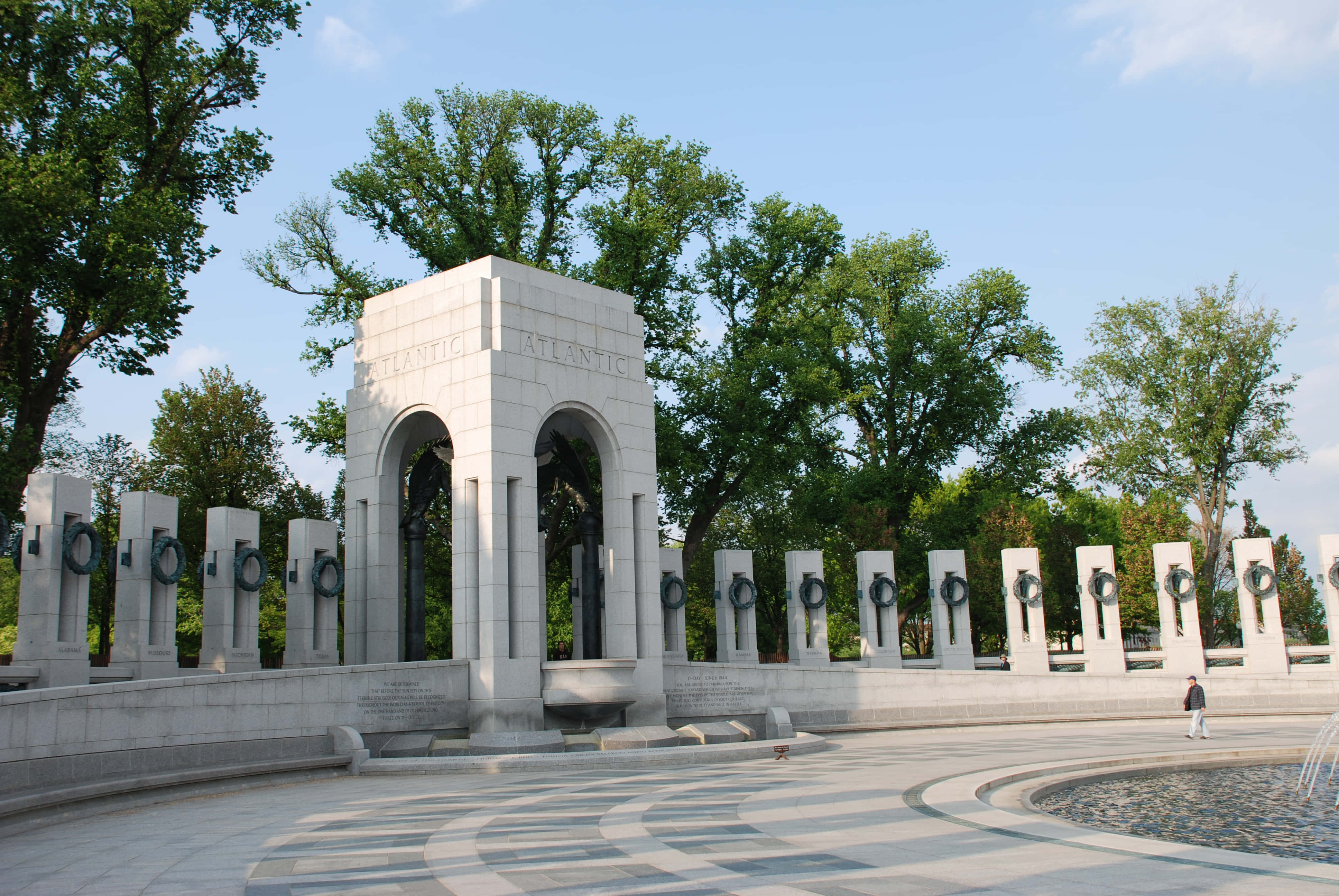 Memorial 桌布 壁紙 — 一座宏偉的白色石頭紀念碑，帶有拱形開口和花圈，矗立在晴朗的藍天下，周圍環繞著茂密的綠樹。
