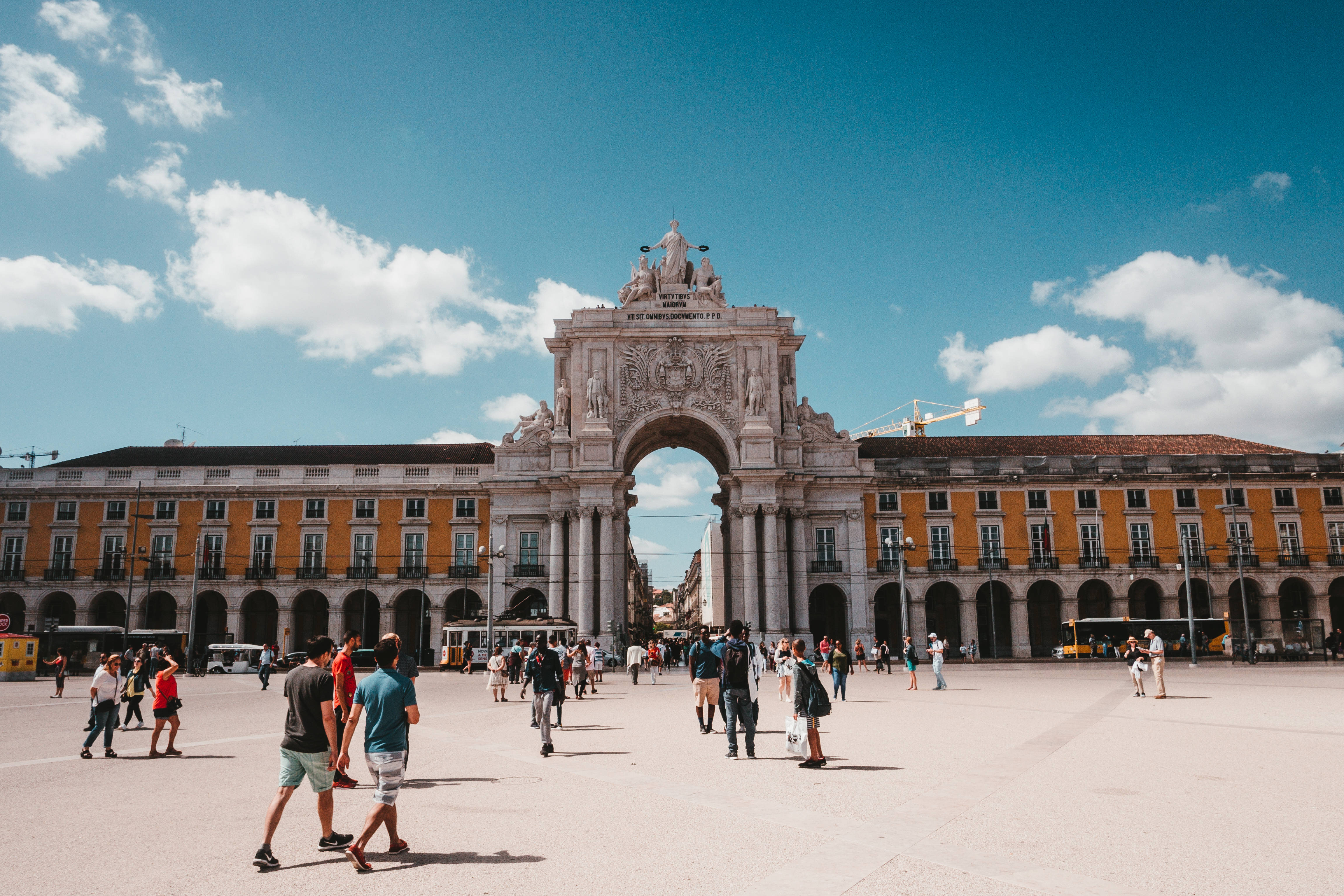 Portugal 桌布 壁紙 — 一座宏偉的石拱門，上面有精緻的雕刻，矗立在陽光普照的廣場上，廣場上滿是在明亮的藍天和白雲下行走的人們。