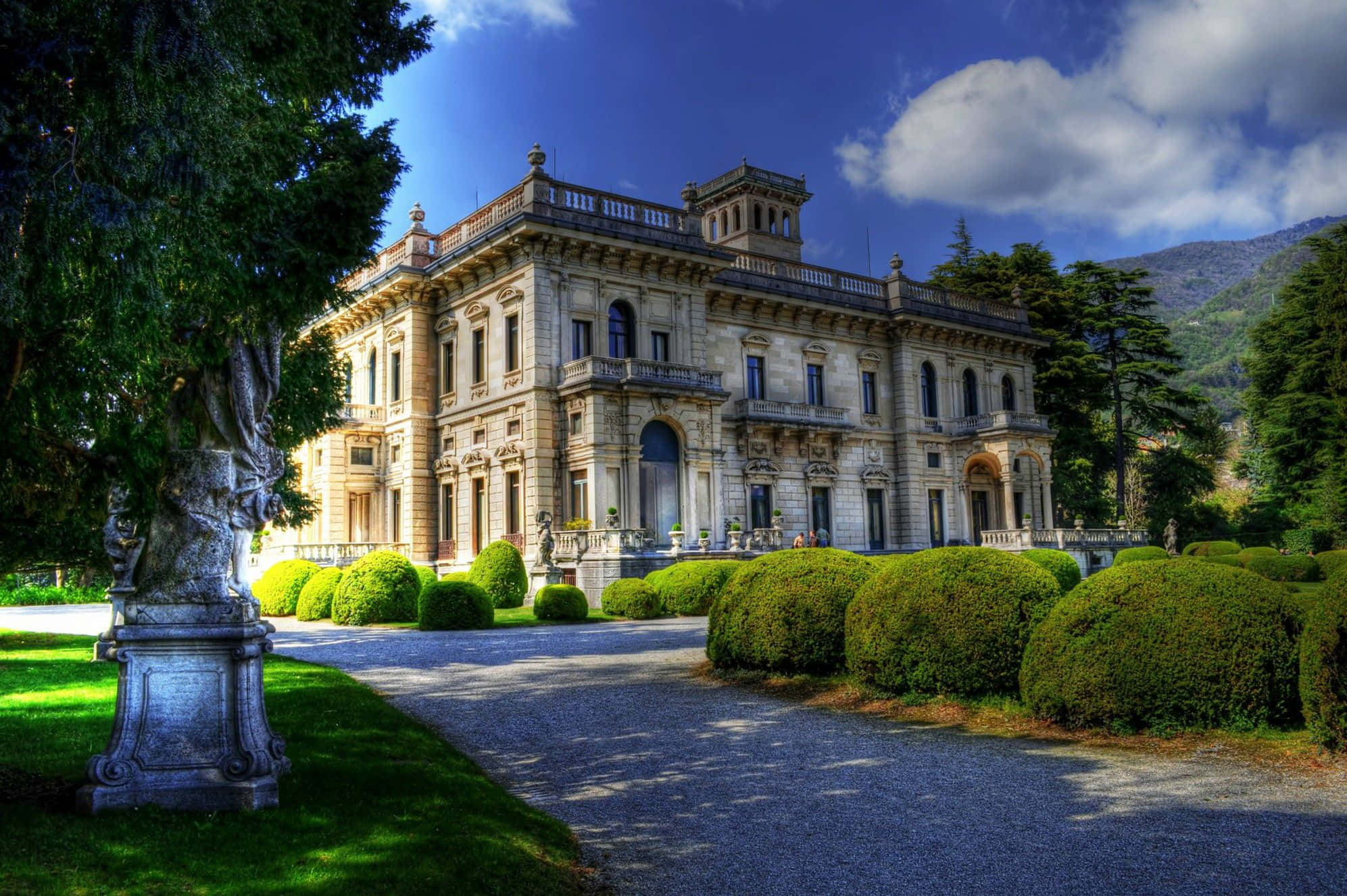 Mansion 桌布 壁紙 — 一座宏偉的石造別墅，帶有華麗的陽台和柱子，矗立在明亮的藍天下，兩旁是修剪整齊的樹籬和碎石車道。