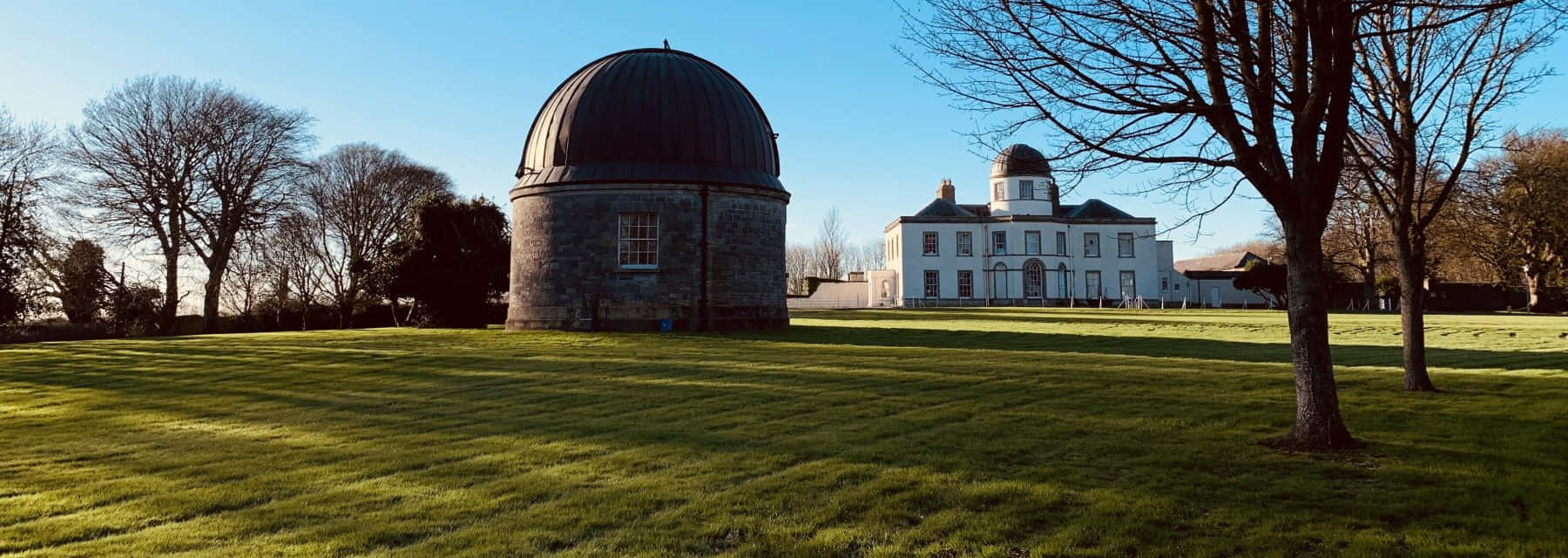 Observatory 桌布 壁紙 — 一座宏偉的石造天文台，帶有深色圓頂，矗立在陽光明媚的草地上，旁邊是一座白色莊園，天空清澈湛藍。