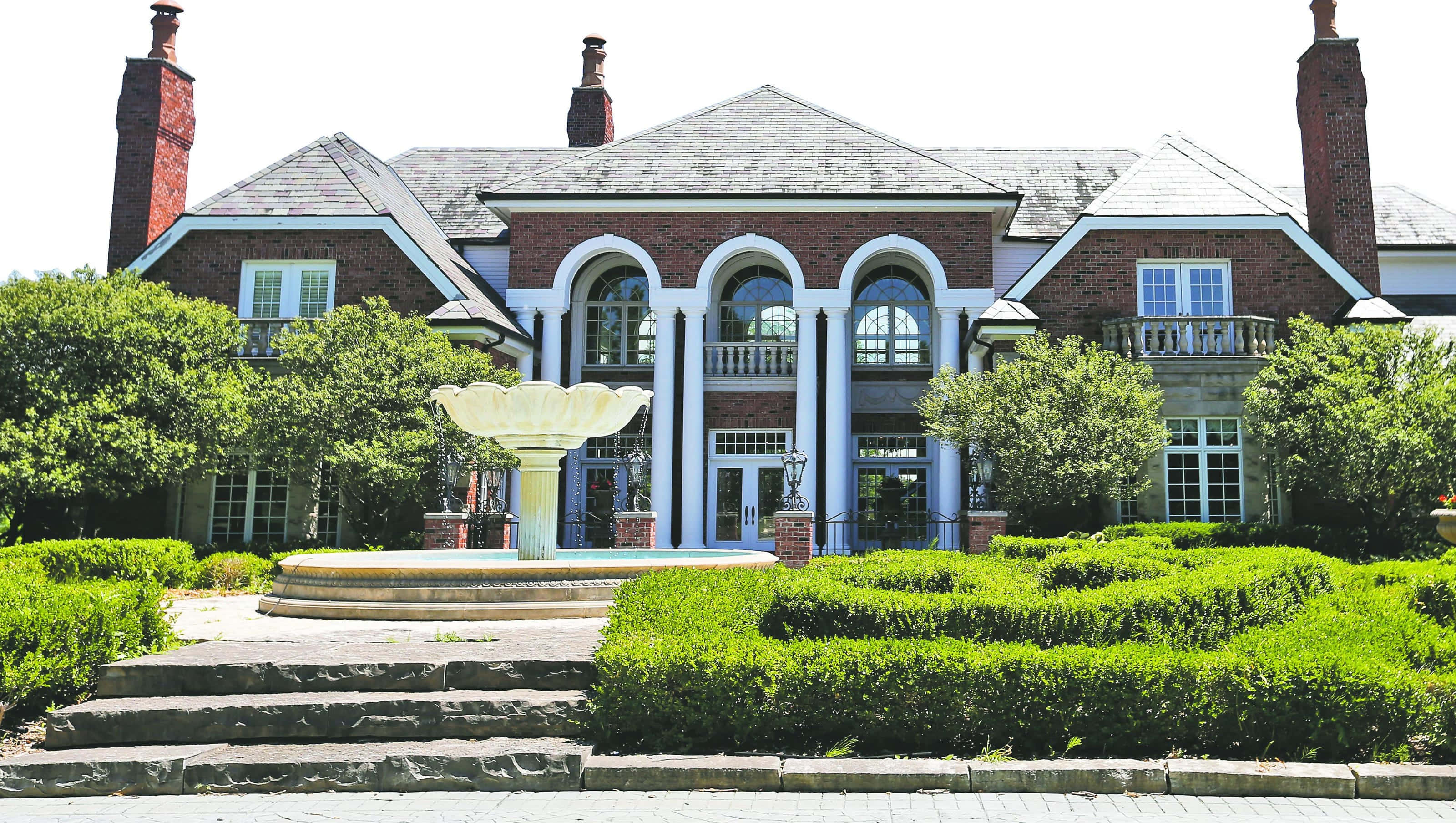 Big House 桌布 壁紙 — 一座宏偉的磚砌豪宅，擁有拱形窗戶和煙囪，矗立在修剪整齊的樹籬和石噴泉後方，沐浴在明亮的日光下。