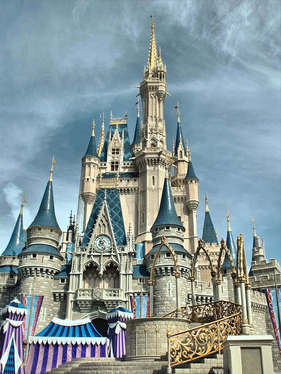 Disney Castle 桌布 壁紙 — 一座宏偉的童話城堡，擁有藍色圓錐形屋頂和金色尖塔，聳立在戲劇性的多雲天空下。