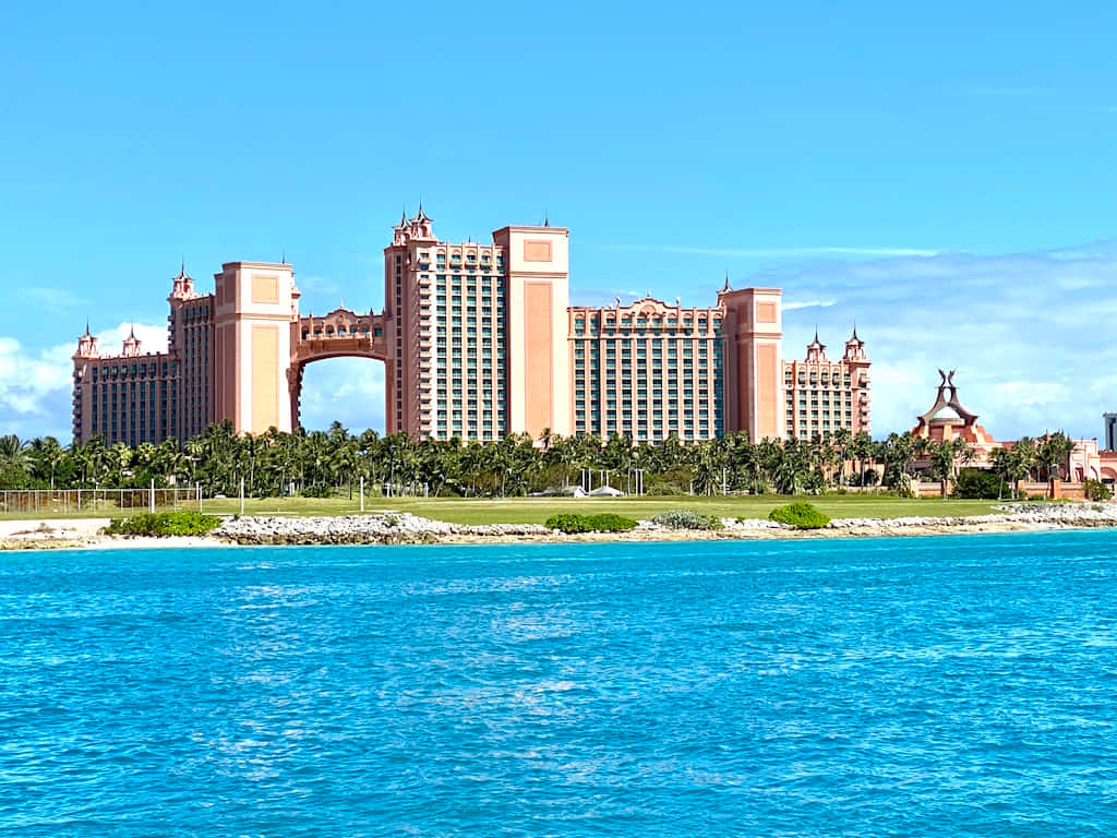 Bahamas Island 桌布 壁紙 — 一座宏偉的粉紅色度假酒店，中央有拱門，雄偉地矗立在熱帶海岸旁，在清澈藍天下，旁邊是碧綠的海水。