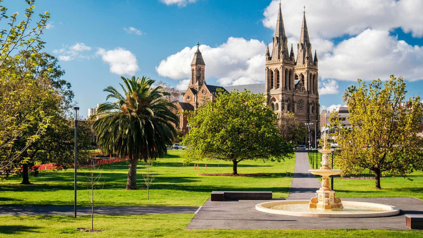 Adelaide 桌布 壁紙 — 一座宏偉的雙尖塔石砌大教堂聳立在陽光明媚的公園後方，公園中央有噴泉、棕櫚樹和生機勃勃的綠草，藍天白雲。