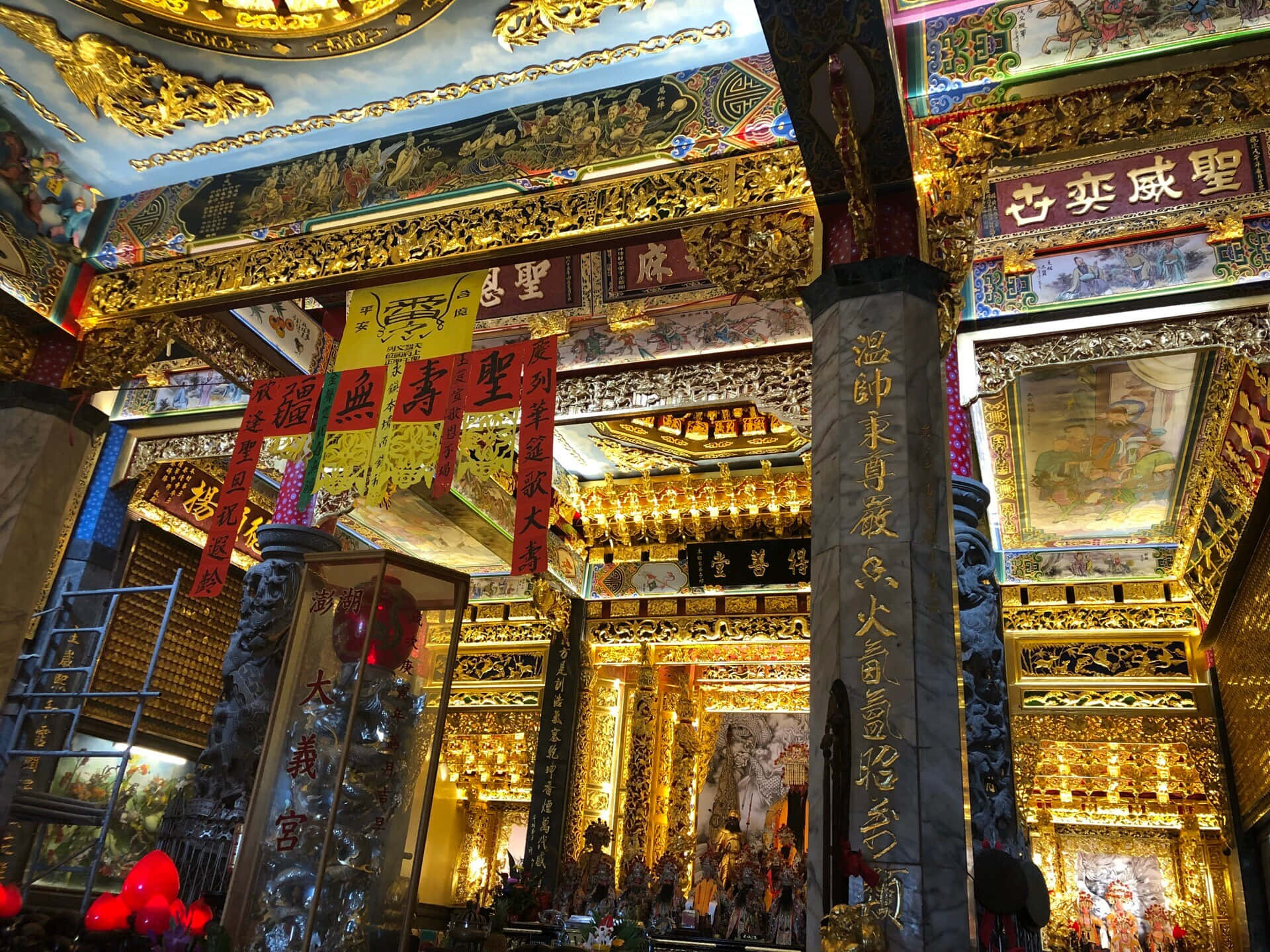 一座寧靜寺廟置身於美麗的風景中，象徵和平與寧靜 壁紙 — 華麗的金色寺廟內部，有精緻的雕刻、紅色橫幅和發光的燈籠，下方是彩繪天花板。