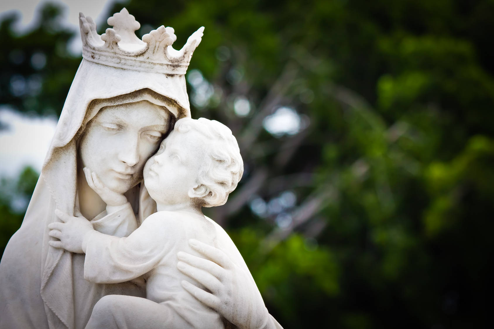 Mary 桌布 壁紙 — 一座寧靜的白色大理石聖母瑪利亞雕像，頭戴冠冕，溫柔地懷抱嬰兒耶穌，背景是柔和的綠色樹葉。