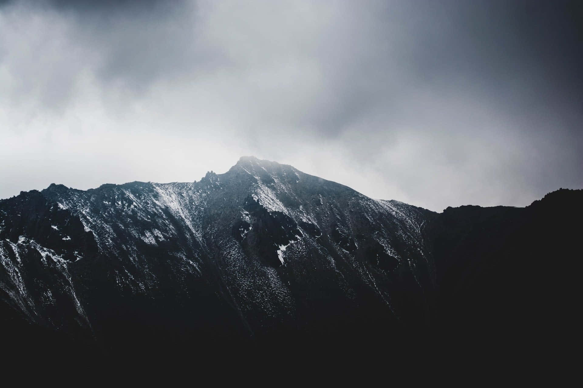 Dark Mountain 桌布 壁紙 — 一座峻峭的山峰刺穿厚重的灰色雲層，積雪附著在崎嶇的山脊上，光線昏暗。