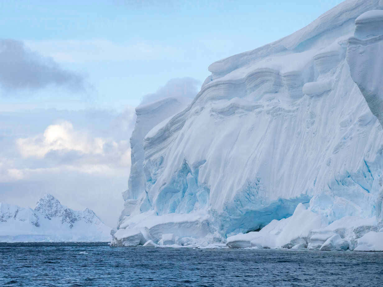 Antarctica 桌布 壁紙 — 一座巨大的冰山，帶有分層的藍白色冰層，聳立在平靜的海面上，天空部分多雲。