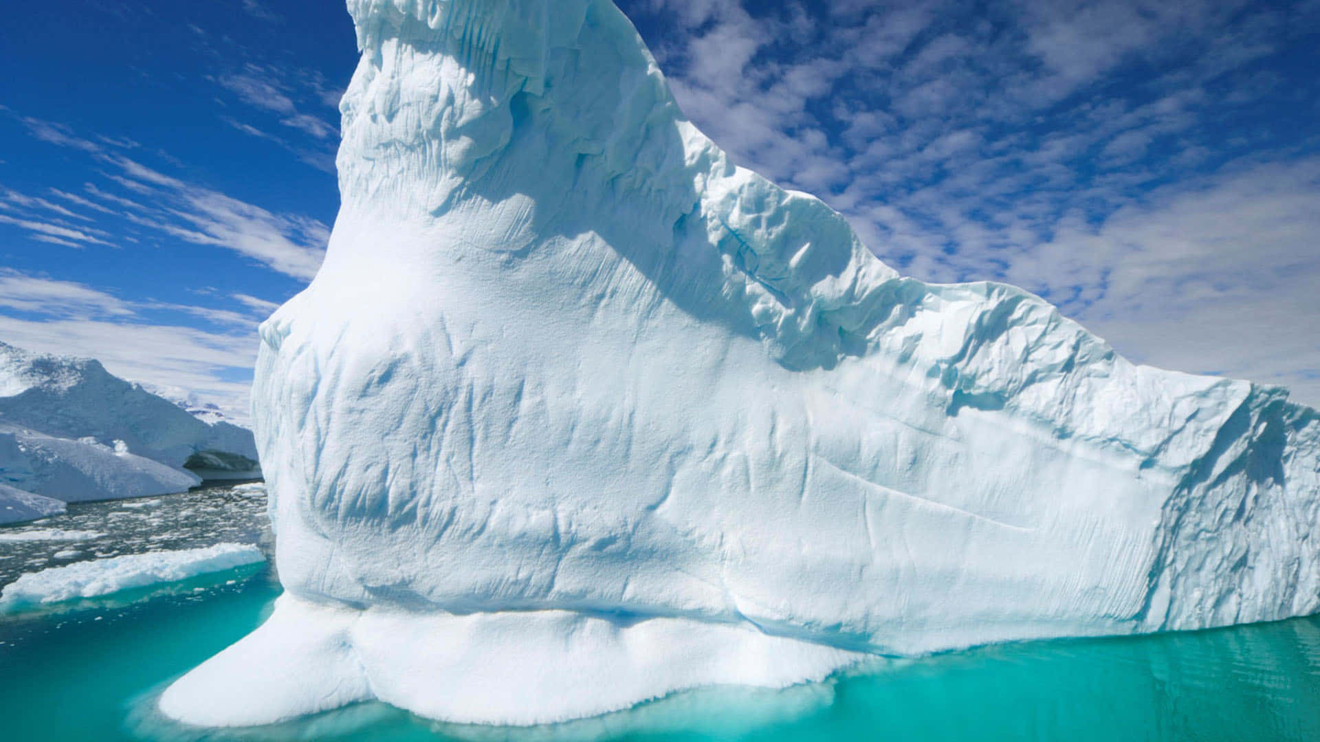 Antarctica 桌布 壁紙 — 一座巨大的冰山，帶有紋理的白色冰塊，漂浮在充滿活力的藍綠色水域中，上方是明亮的藍天，點綴著幾朵雲。