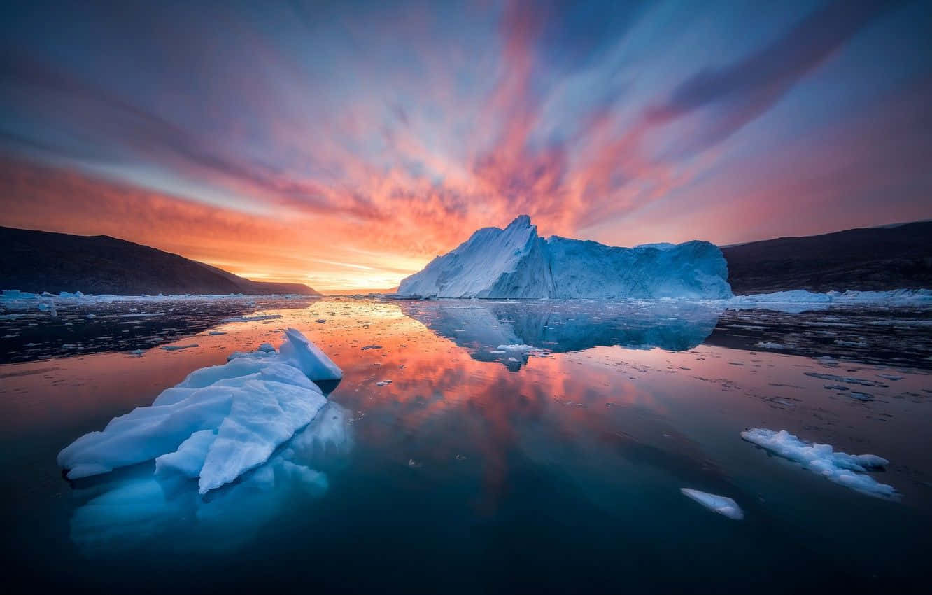 Greenland 桌布 壁紙 — 一座巨大的冰山在平靜的北極海面上反射出鮮豔的粉紅色和橙色日落雲彩，周圍有較小的浮冰。
