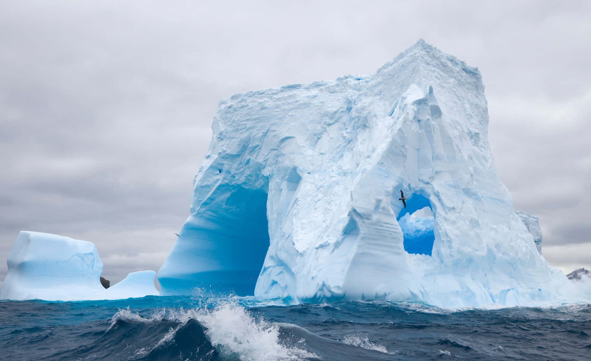 Antarctica 桌布 壁紙 — 一座巨大的冰山帶有天然拱門，在波濤洶湧的藍色海水中聳立，天空多雲。