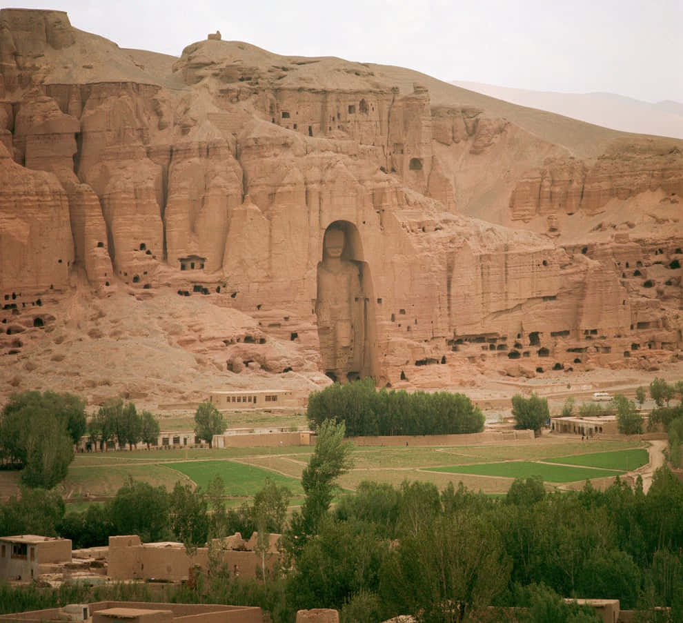 Afghanistan 桌布 壁紙 — 一座巨大的古代佛像雕刻在砂岩懸崖上，俯瞰著朦朧天空下的綠色農田和小型建築。