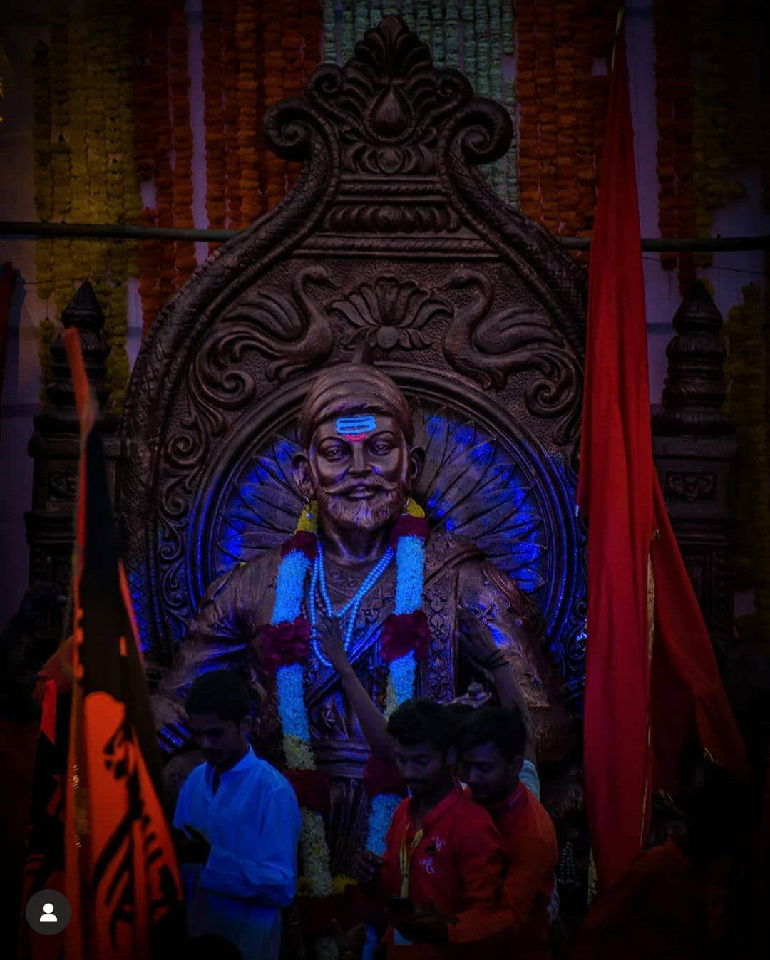 Indian 桌布 壁紙 — 一座巨大的照明神像，鬍鬚神祇裝飾著花環和紅旗，在寺廟環境中被信徒圍繞。