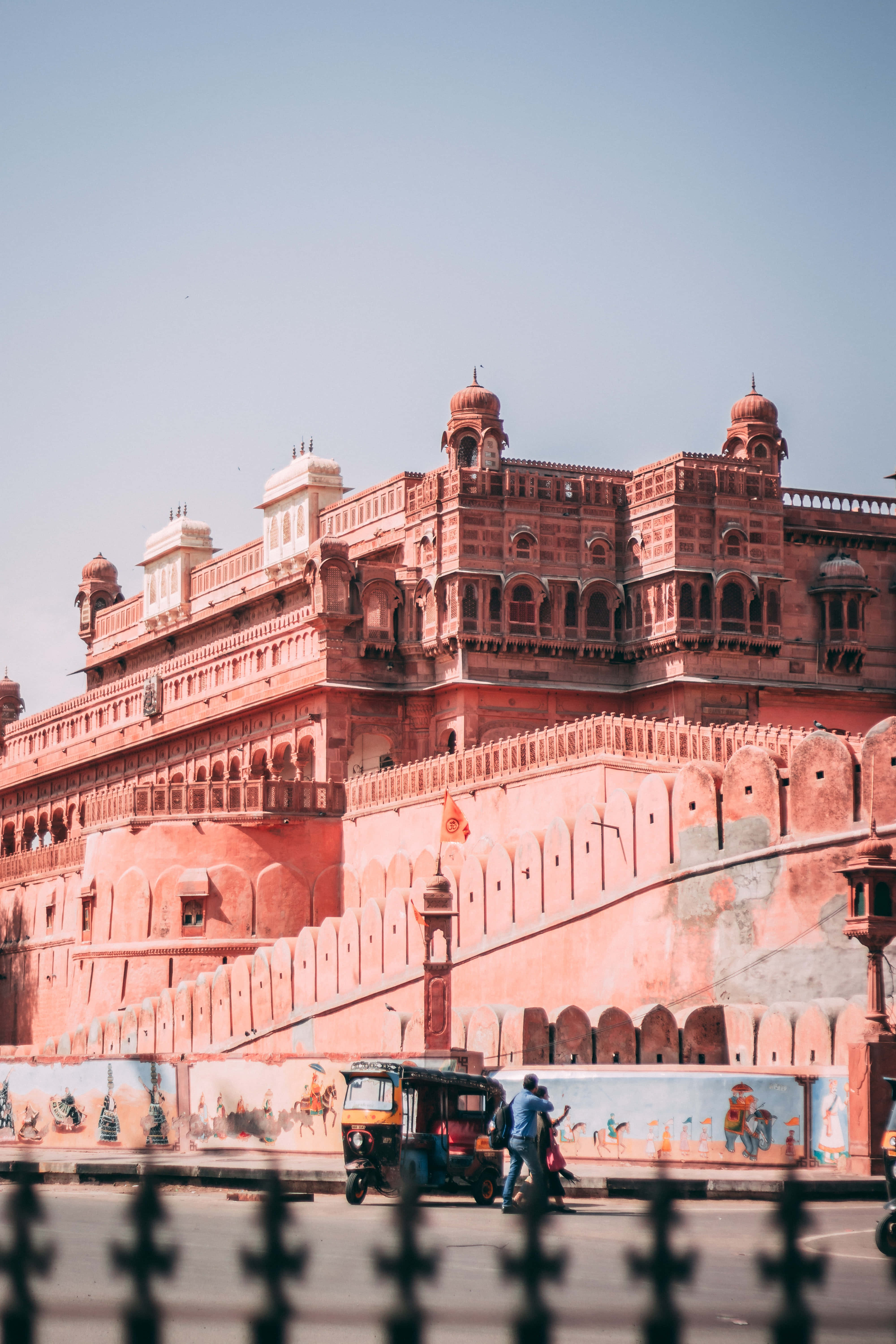 Rajasthan 桌布 壁紙 — 一座巨大的粉紅色砂岩堡壘，帶有圓頂和拱門，聳立在一條街道上，一輛人力車在晴朗的藍天下經過。