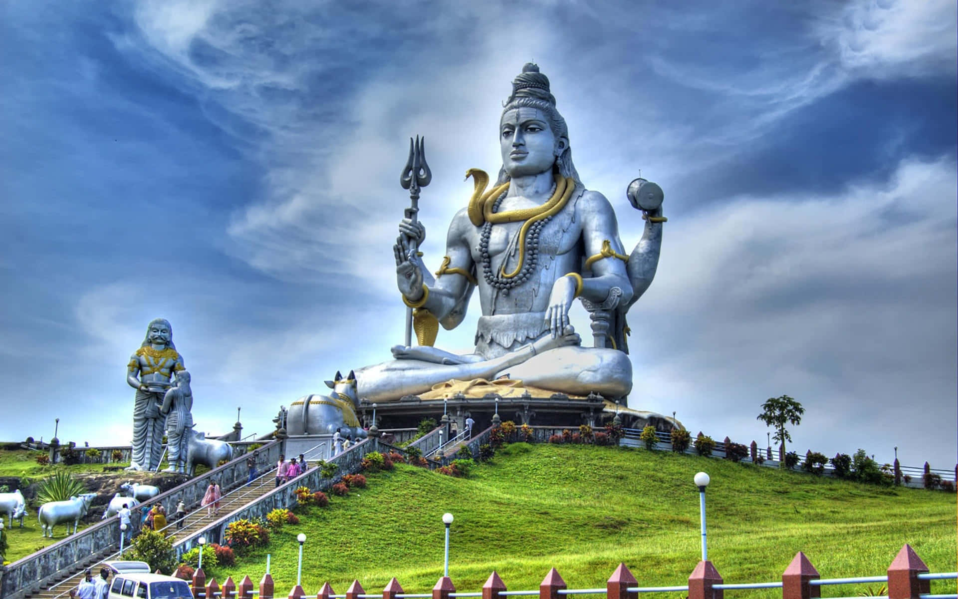 Shiva 桌布 壁紙 — 一座巨大的銀色濕婆神像在戲劇性的藍天白雲下，寧靜地坐落在翠綠的山丘頂上。