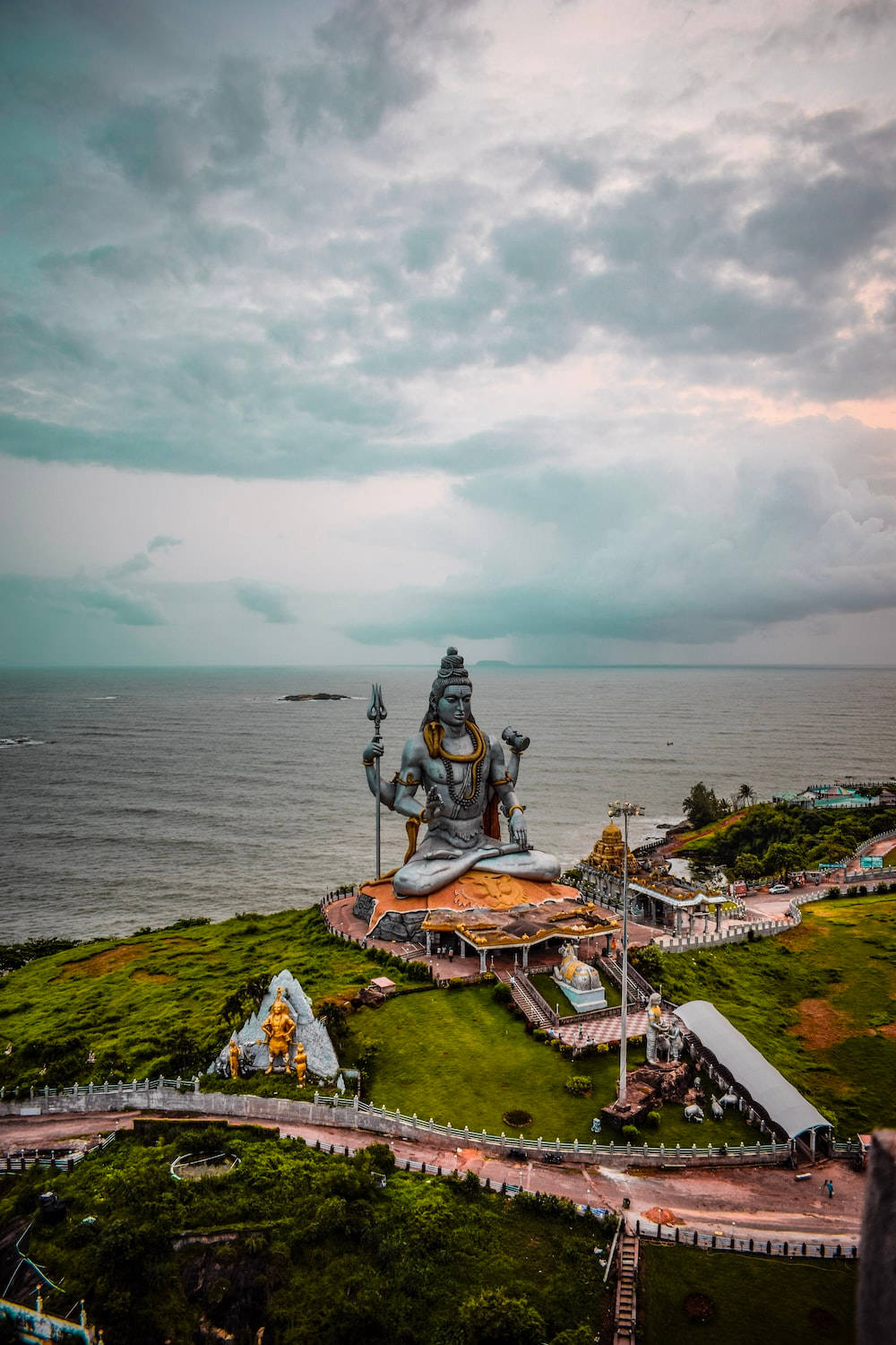 India 桌布 壁紙 — 一座巨大的銀色濕婆神像寧靜地坐在鬱鬱蔥蔥的綠色懸崖上，俯瞰著廣闊的海洋，天空佈滿戲劇性的雲層。