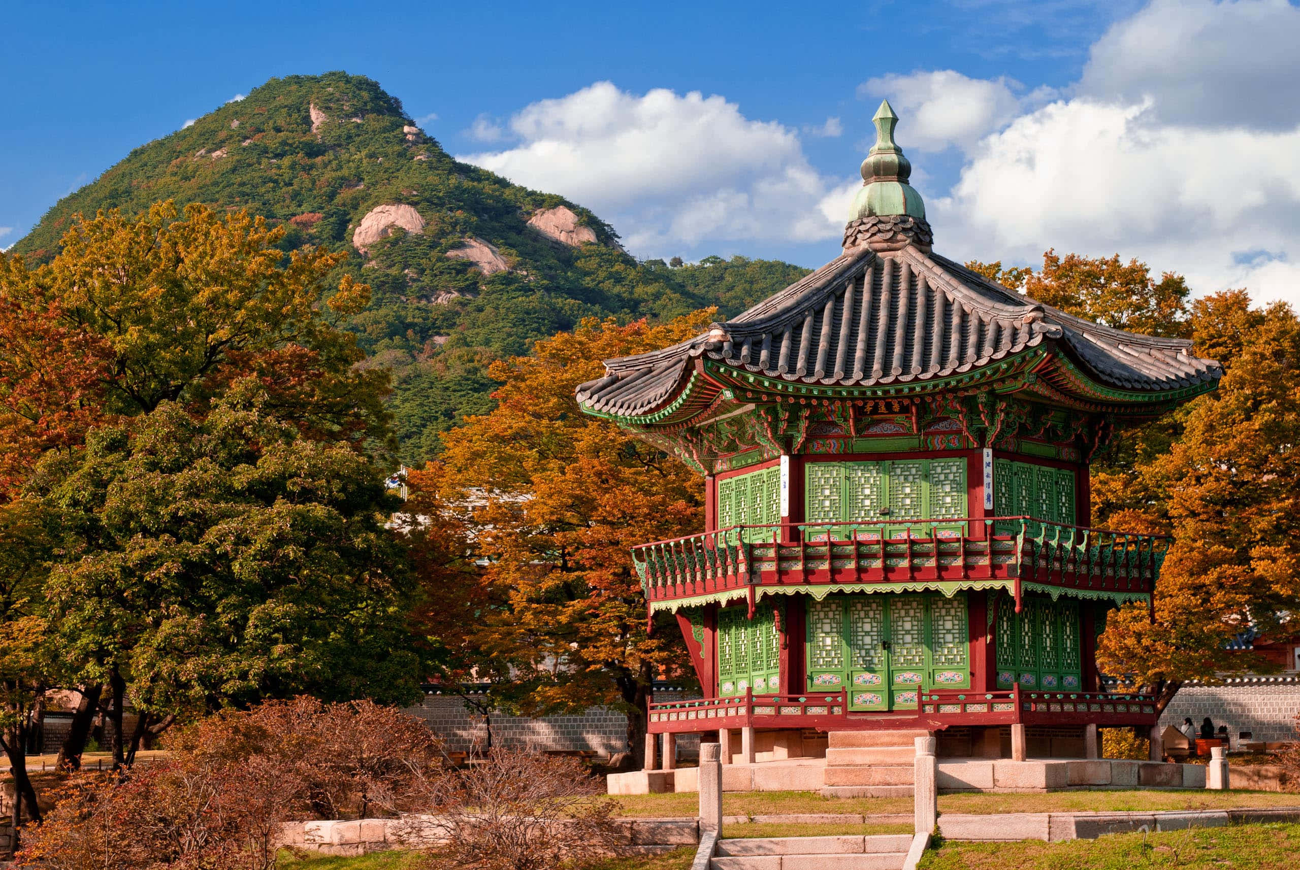 South Korea 桌布 壁紙 — 一座帶有綠色和紅色點綴的傳統韓國亭閣矗立在秋樹之中，背景是森林覆蓋的山脈，天空湛藍明亮。
