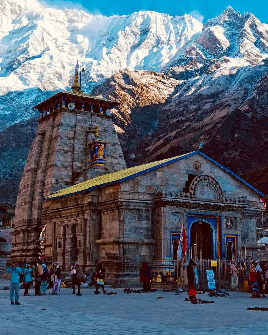 Kedarnath 桌布 壁紙 — 一座帶有藍色和金色點綴的石廟矗立在晴朗藍天下高聳的雪山前。