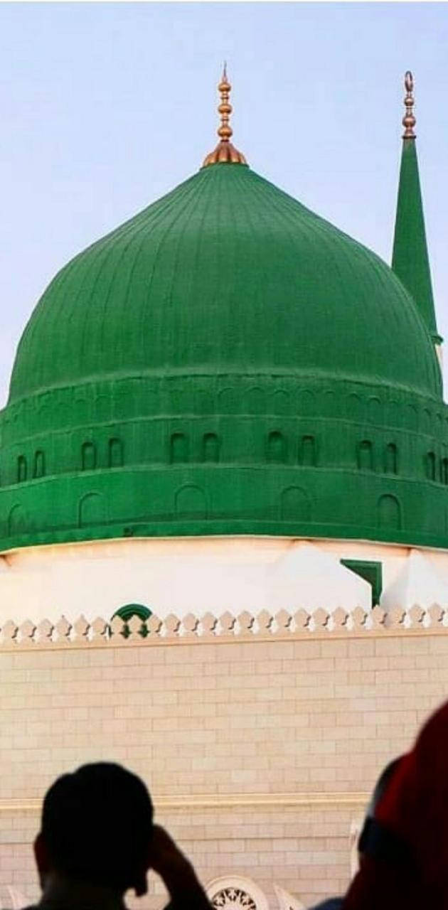 Madina Sharif 桌布 壁紙 — 一座帶有金色尖頂的大型綠色圓頂清真寺矗立在晴朗的天空下，剪影般的信徒們抬頭仰望。