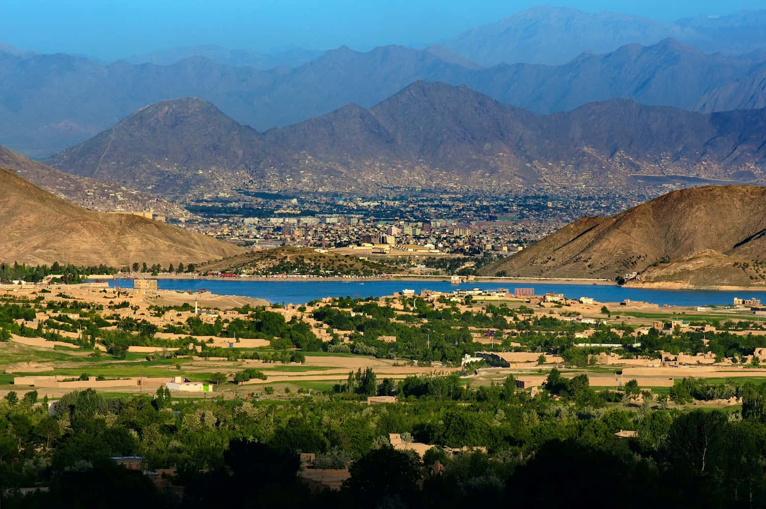 Afghanistan 桌布 壁紙 — 一座廣闊的城市坐落在乾燥的山丘和清澈天空下充滿活力的藍色湖泊之間。