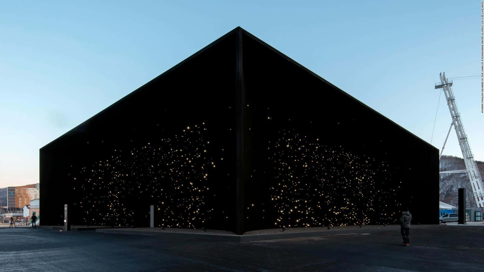 Vantablack 桌布 壁紙 — 一座引人注目的黑色棱角建築，外牆嵌有發光的燈光，在晴朗的藍天下矗立，附近有人走動。