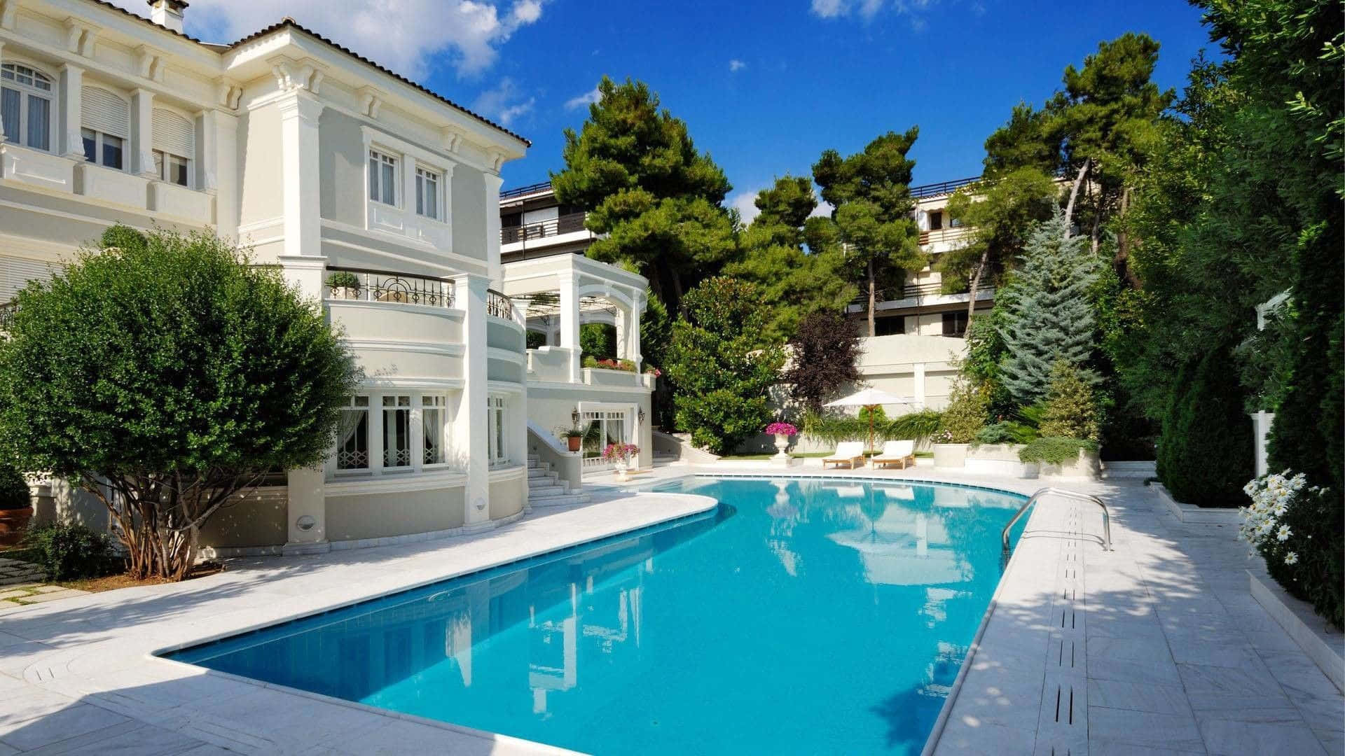 Mansion 桌布 壁紙 — 一座擁有古典建築風格的大型白色別墅俯瞰著一個水晶藍色的游泳池，周圍環繞著茂密的綠樹，上方是明亮的藍天。