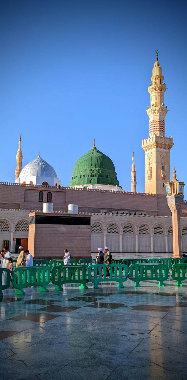 Madina Sharif 桌布 壁紙 — 一座擁有綠色和白色圓頂及高聳宣禮塔的大清真寺矗立在晴朗的藍天下，遊客在綠色屏障附近行走。
