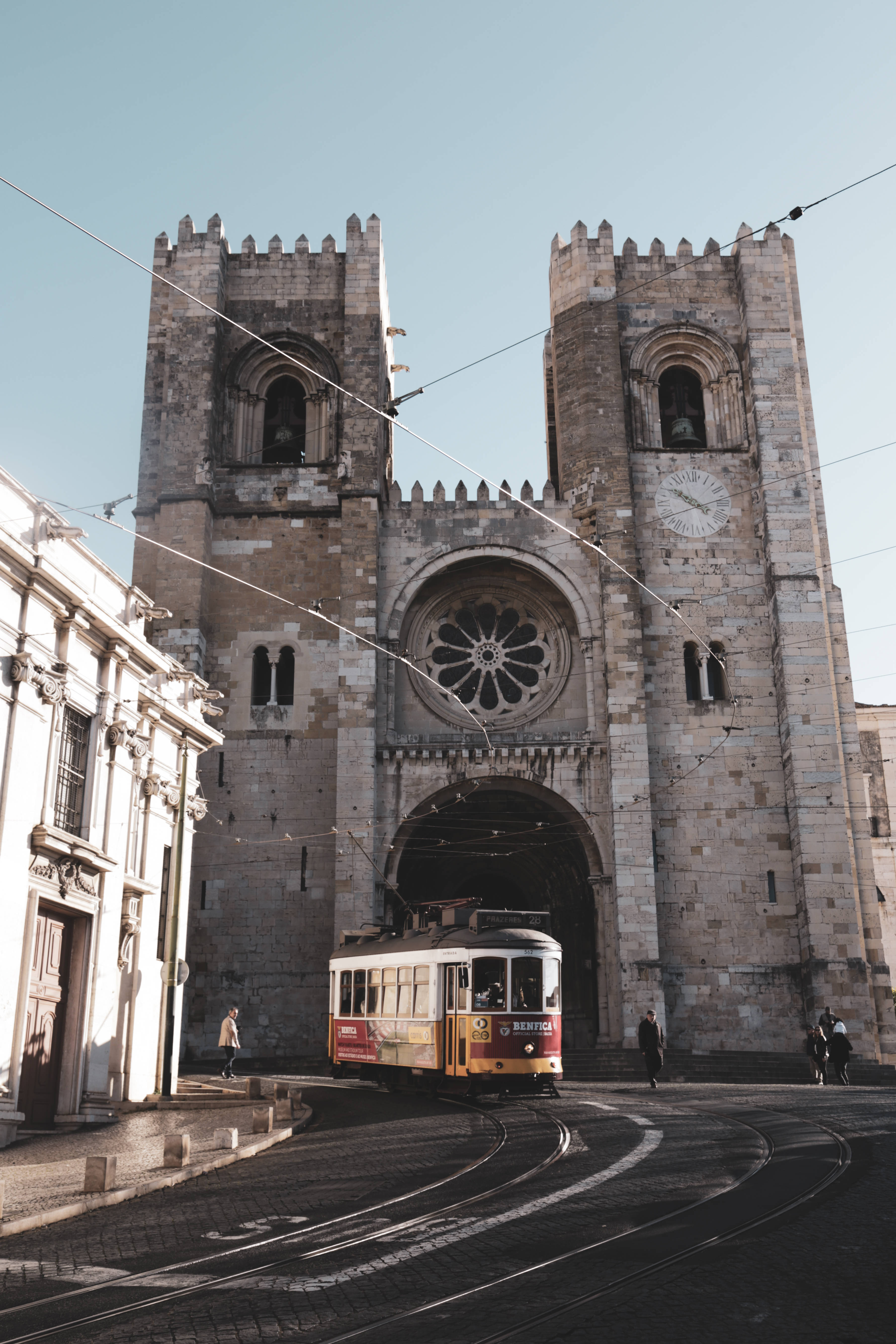 Portugal 桌布 壁紙 — 一座擁有雙塔和玫瑰窗的歷史石造大教堂高聳矗立，一輛復古黃色電車在陽光普照的鵝卵石街道上穿過其拱門。