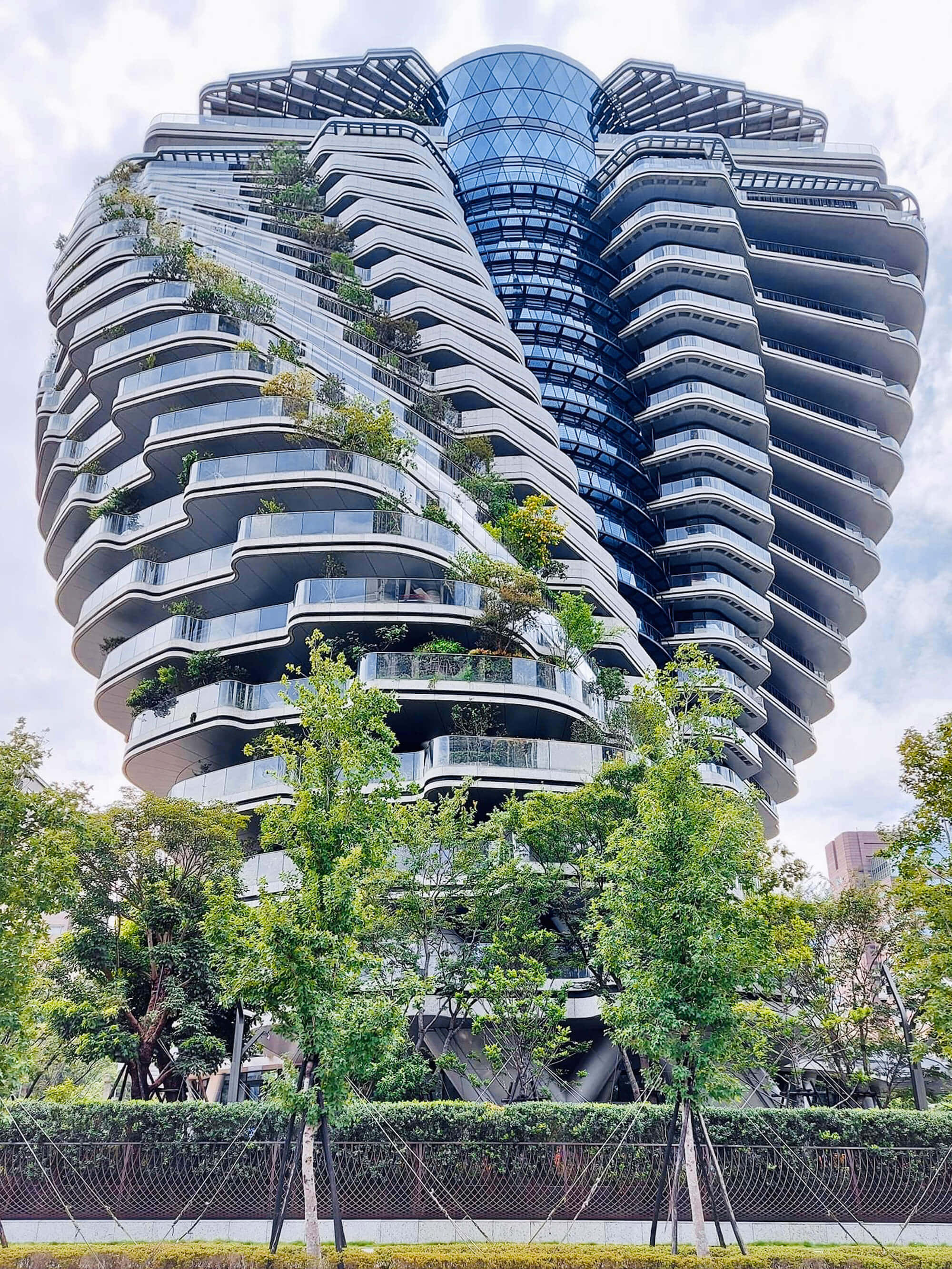 Beautiful Architecture 桌布 壁紙 — 一座未來主義的玻璃摩天大樓，帶有弧形陽台和茂密的綠植，聳立在多雲的天空下。